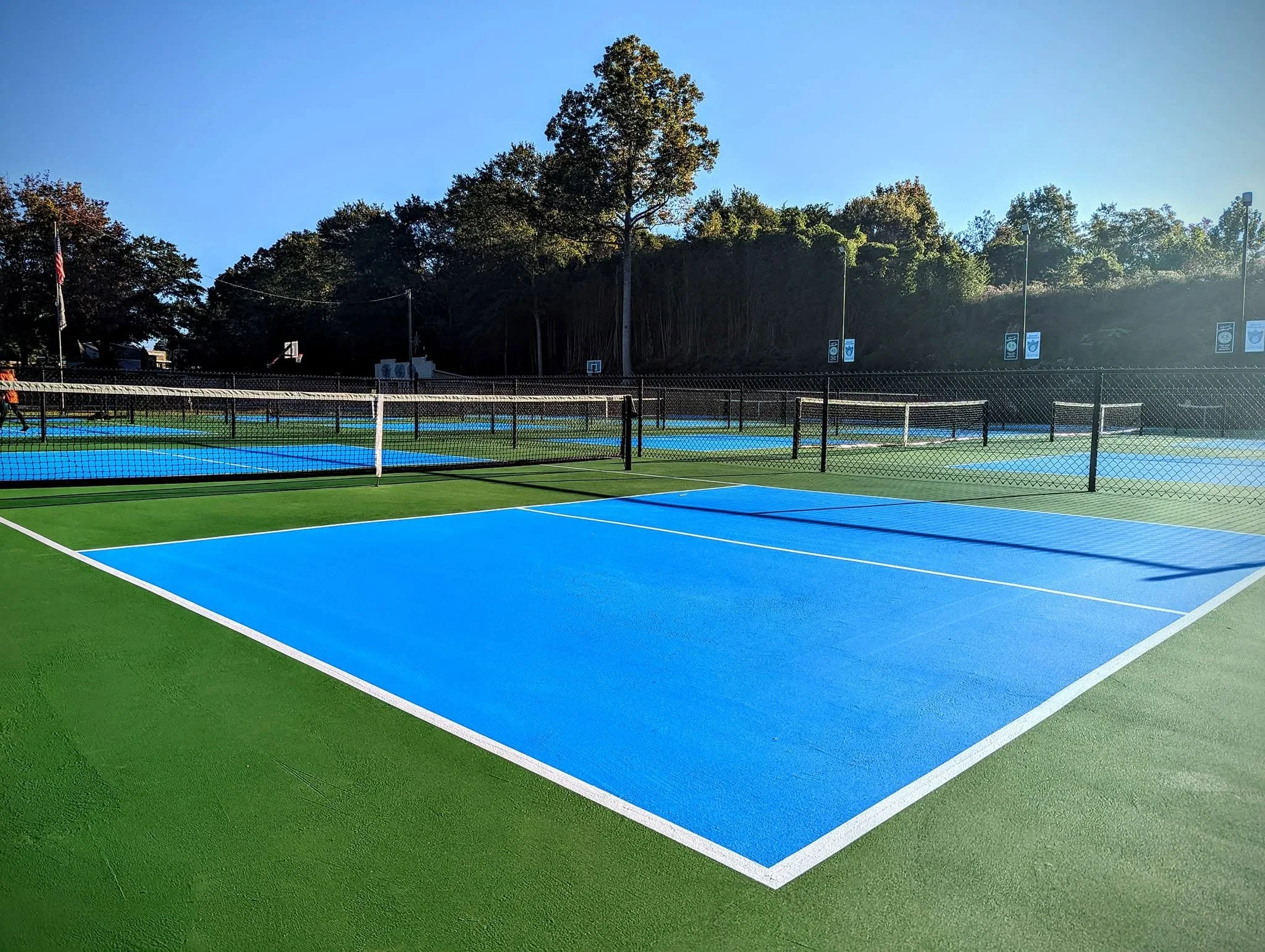 Outdoor tennis courts with artificial green and blue surfaces, surrounded by fencing, with a wooded hillside in the background under a clear blue sky.