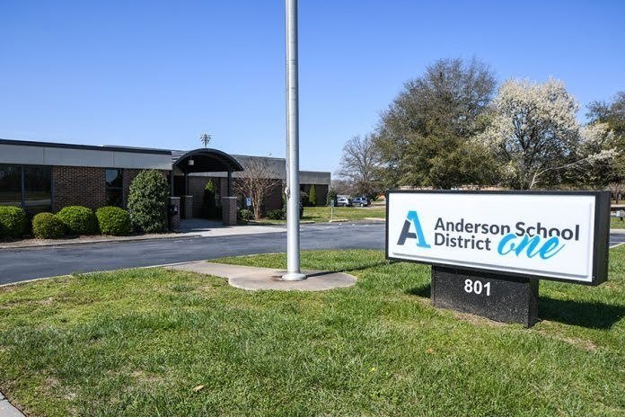 A sign for Anderson School District One in front of a school building with trees and a parking lot under a clear blue sky.