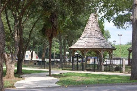 A park with tall trees, a wooden gazebo with a shingled roof, and a paved walking path.