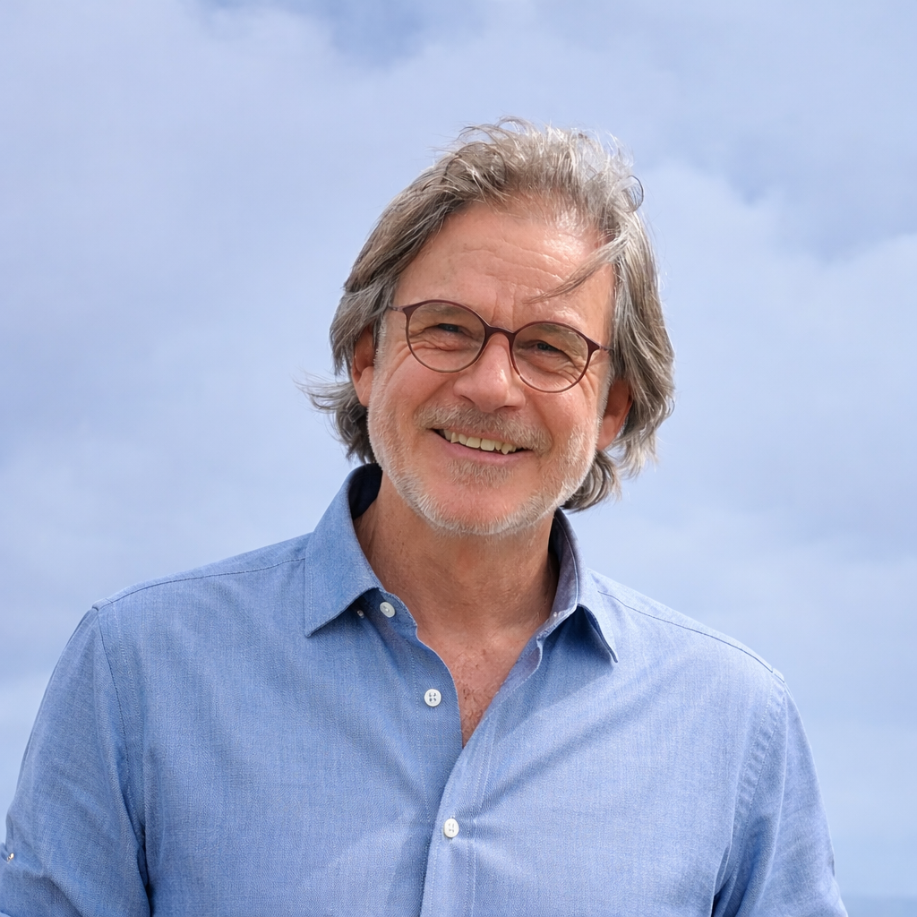Hombre mayor con cabello gris, barba y gafas, sonriendo, usando una camisa azul, de pie al aire libre con cielo nublado al fondo.