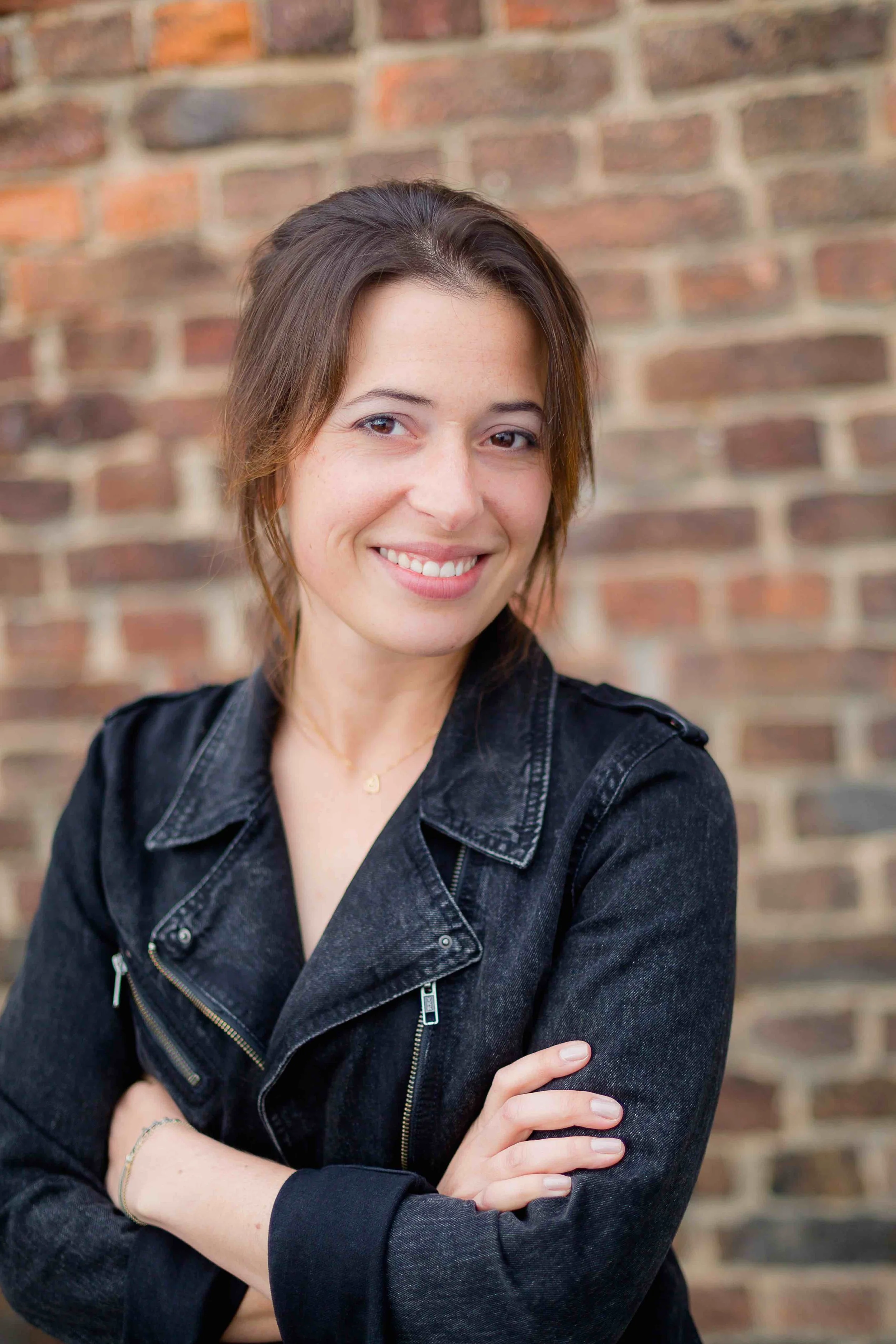 Junge Frau mit braunen Haaren, lächelnd, trägt eine schwarze Jeansjacke, steht vor einer Backsteinmauer.