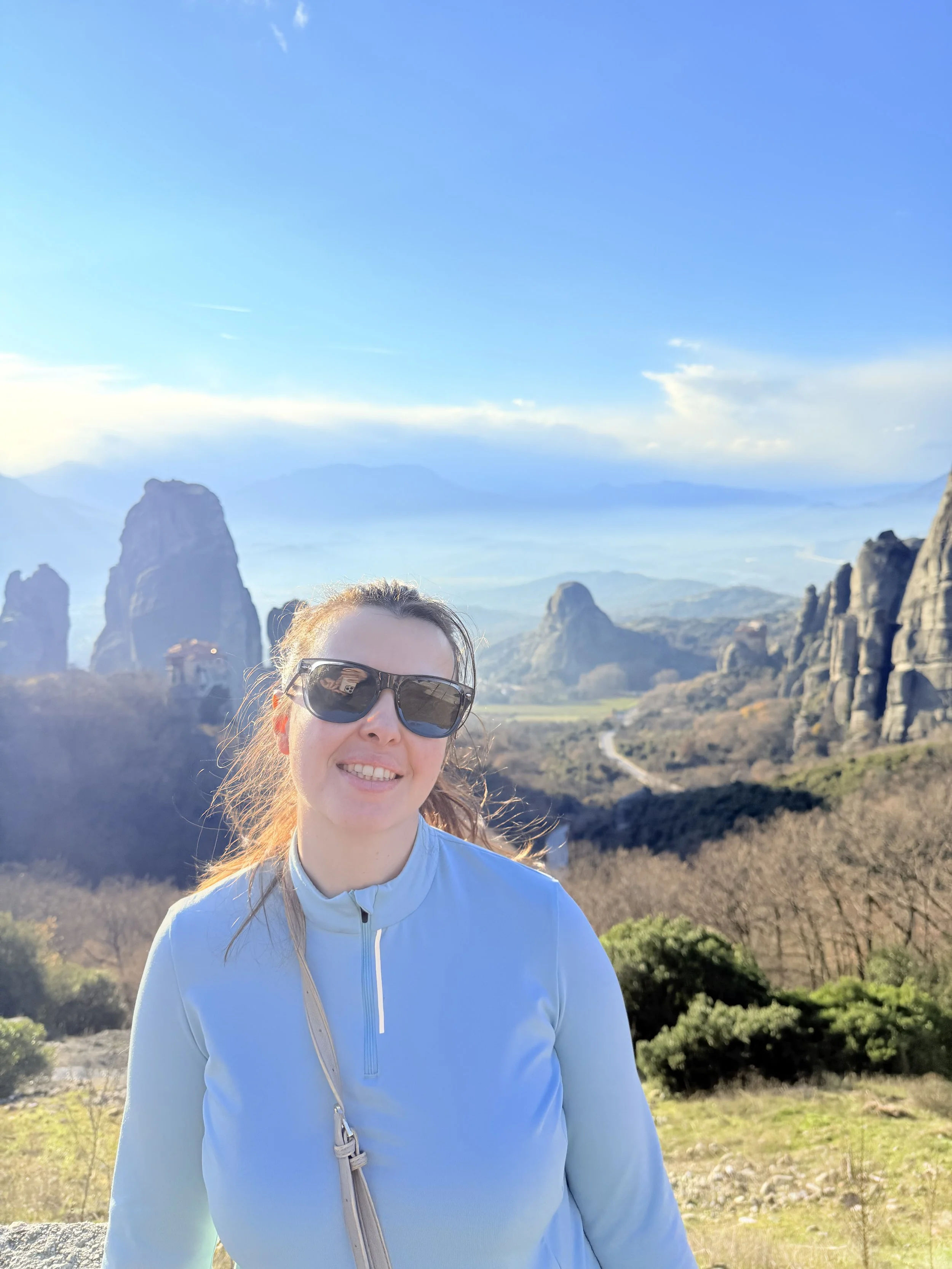 Femme souriante portant des lunettes de soleil et une veste bleue, posant avec un paysage de montagnes rocheuses et un ciel bleu en arrière-plan.