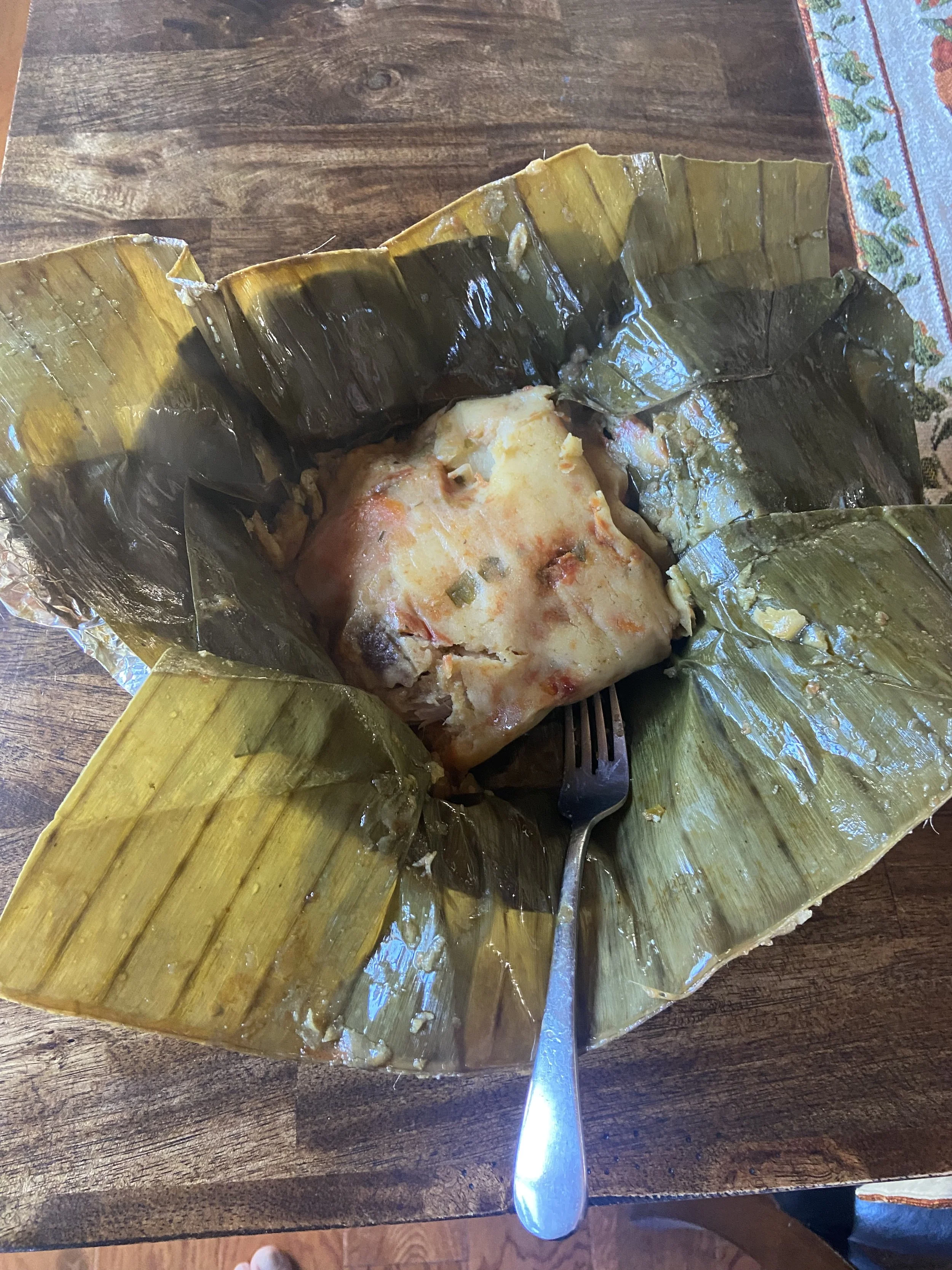 A tamale wrapped in banana leaves with some filling visible, served on a wooden table with a fork.