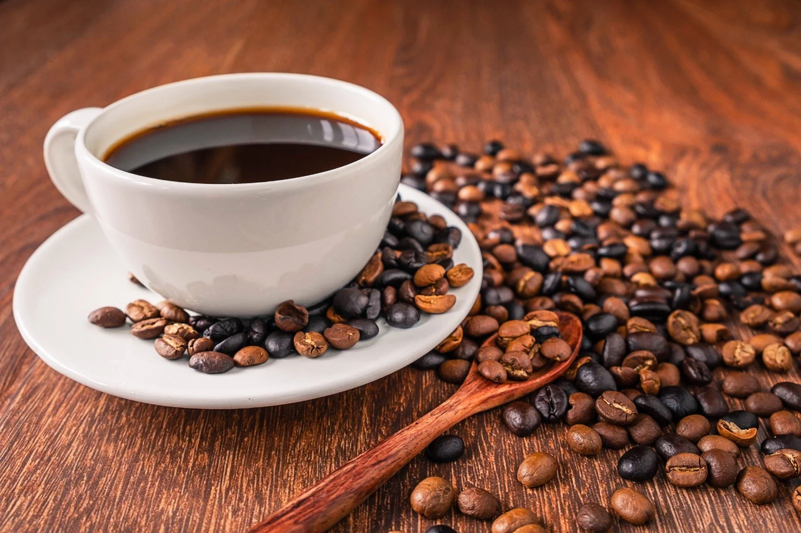 A white coffee cup filled with black coffee on a small white saucer, surrounded by scattered coffee beans on a wooden surface, with a wooden spoon among the beans.