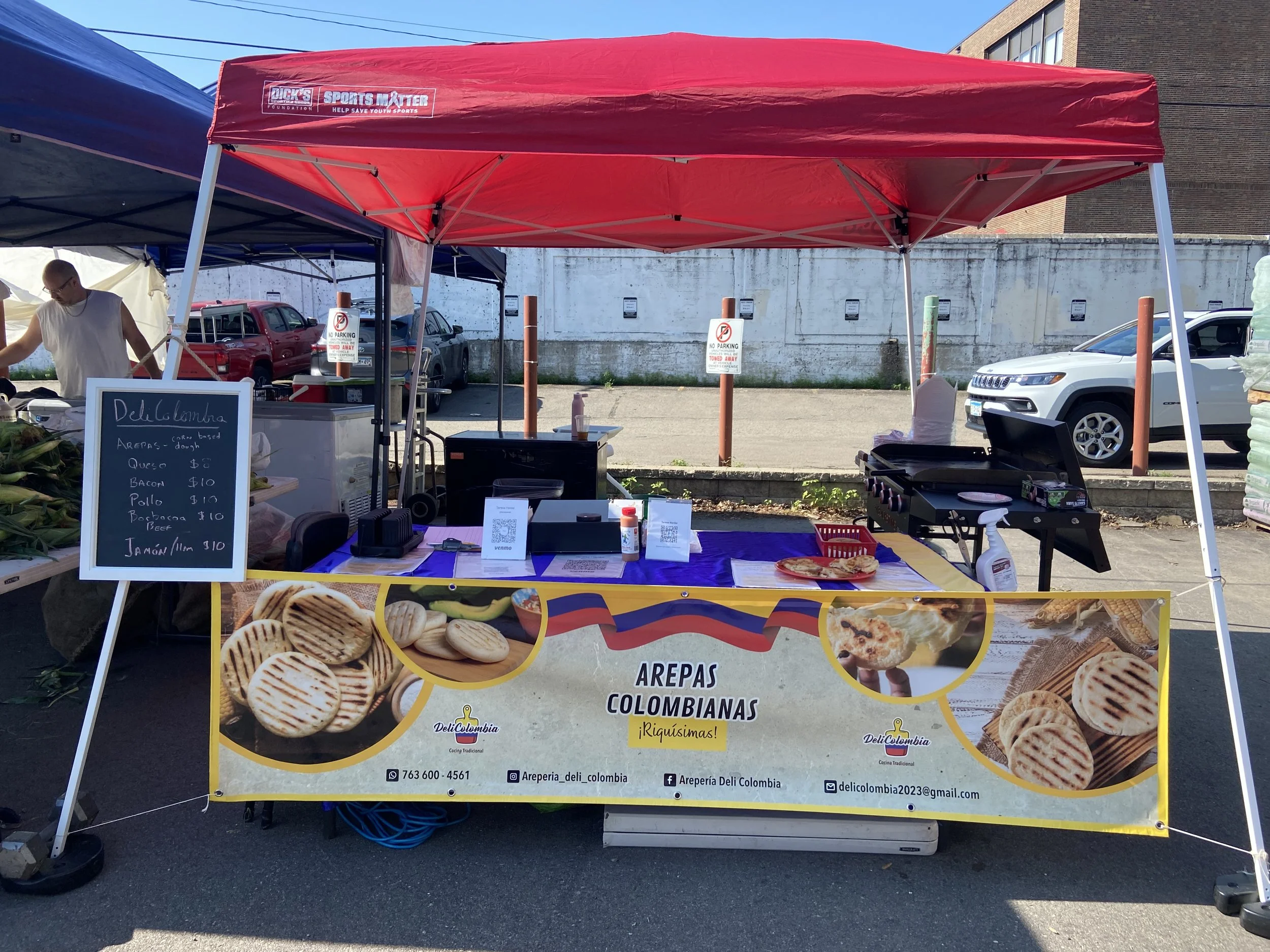 A food stand at an outdoor market with a red canopy selling Colombian arepas. The front banner displays images of arepas and descriptions, including the phrase "Arepas Colombianas". A small chalkboard sign lists ingredients and prices. There is a gri