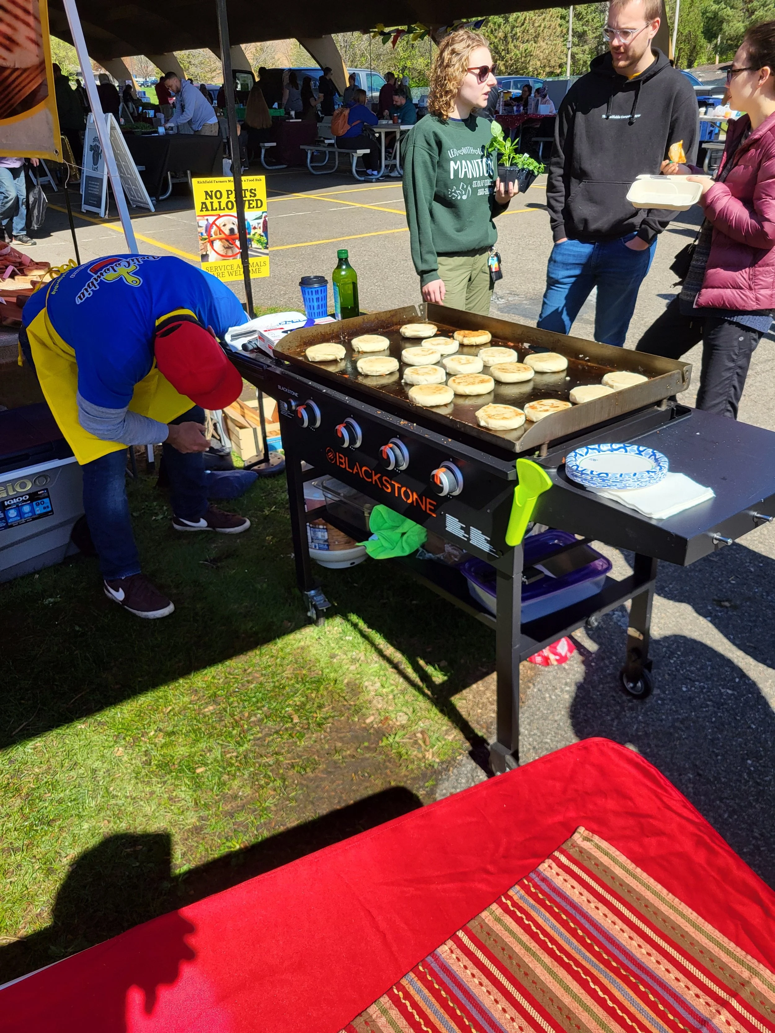 People standing around a grill at an outdoor event, cooking and serving flatbread or pita bread. There are tables and tents in the background with more people.