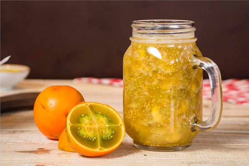 A mason jar filled with a yellowish, bubbling fermented drink with lemon slices, placed on a wooden surface next to whole and halved yellow tomatoes.