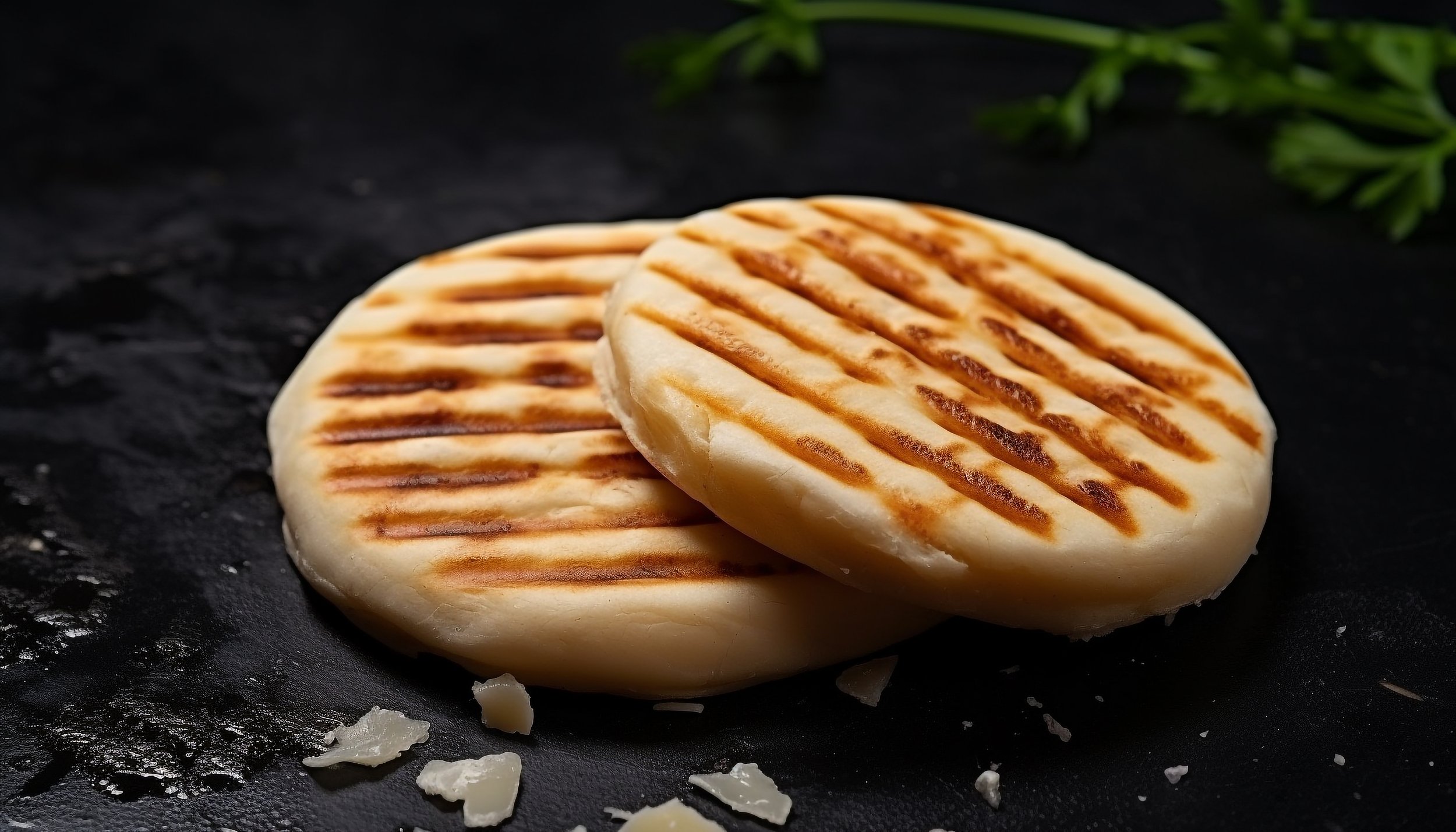 Two grilled Arepas with grill marks on a black surface with some broken pieces of cheese around, and green herbs in the background.