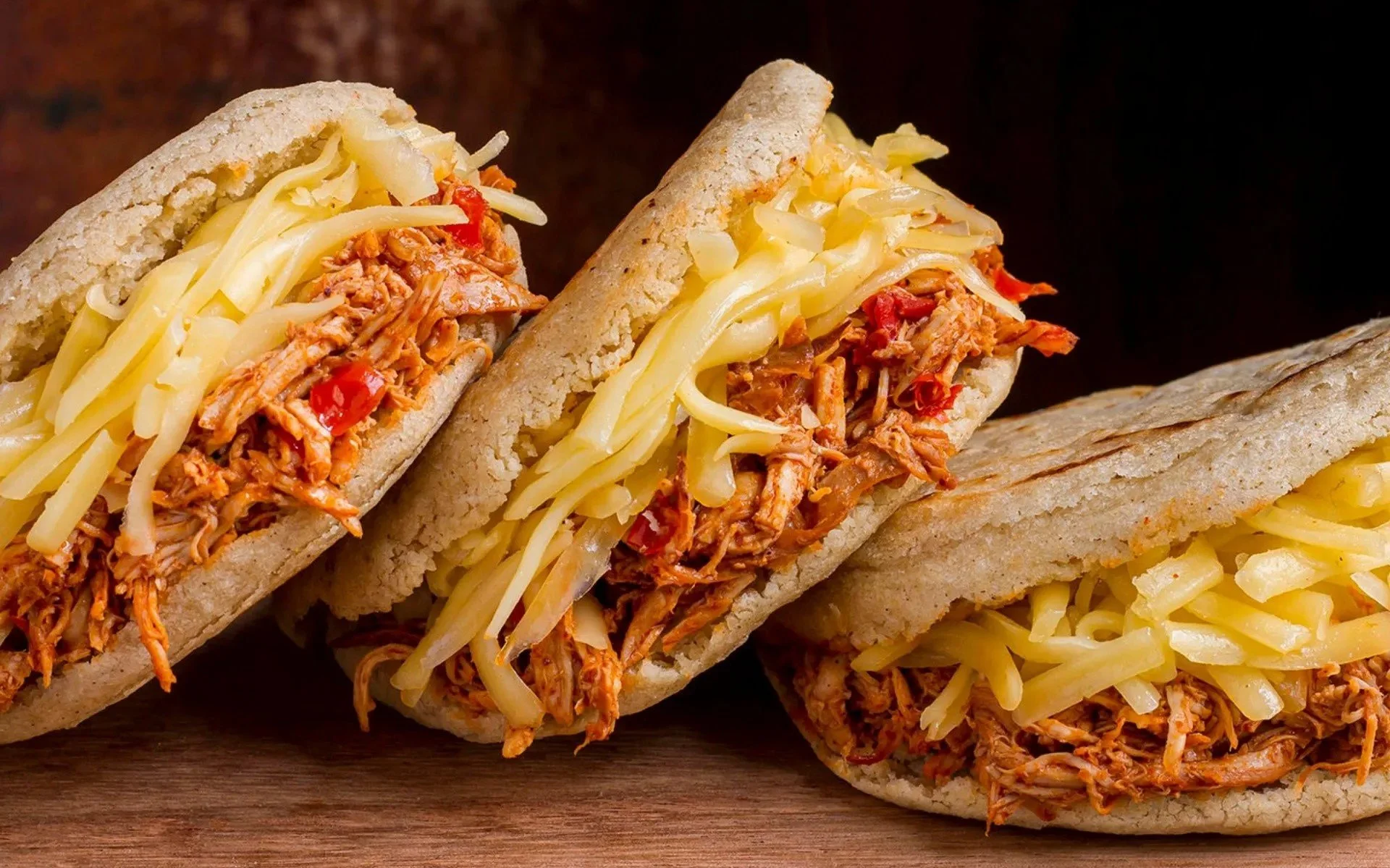 Three shredded chicken sandwiches with cheese in sandwich buns on a wooden surface.