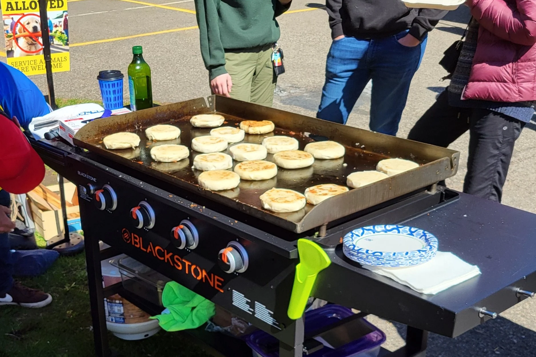 A Blackstone griddle with arepas cooking on it, set up outdoors. Several people stand around, observing. There are paper plates, napkins, and various beverage bottles nearby, with a sign in the background.