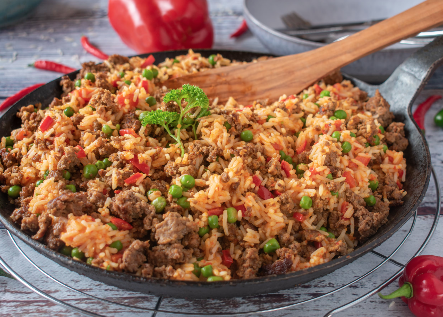 Heavily loaded skillet of seasoned ground beef, rice, peas, and red bell peppers garnished with a parsley sprig, with a wooden spatula inside and chili peppers around it.