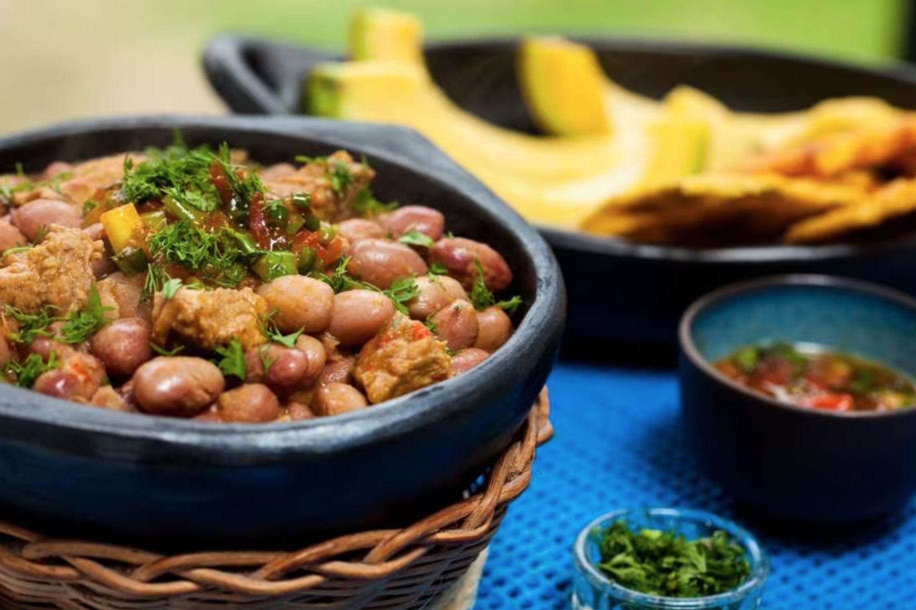 A traditional Colombian meal featuring a bowl of beans topped with cilantro, a small bowl of salsa, another bowl of sauce, a bowl of chopped cilantro, and plates of sliced and fried plantains.