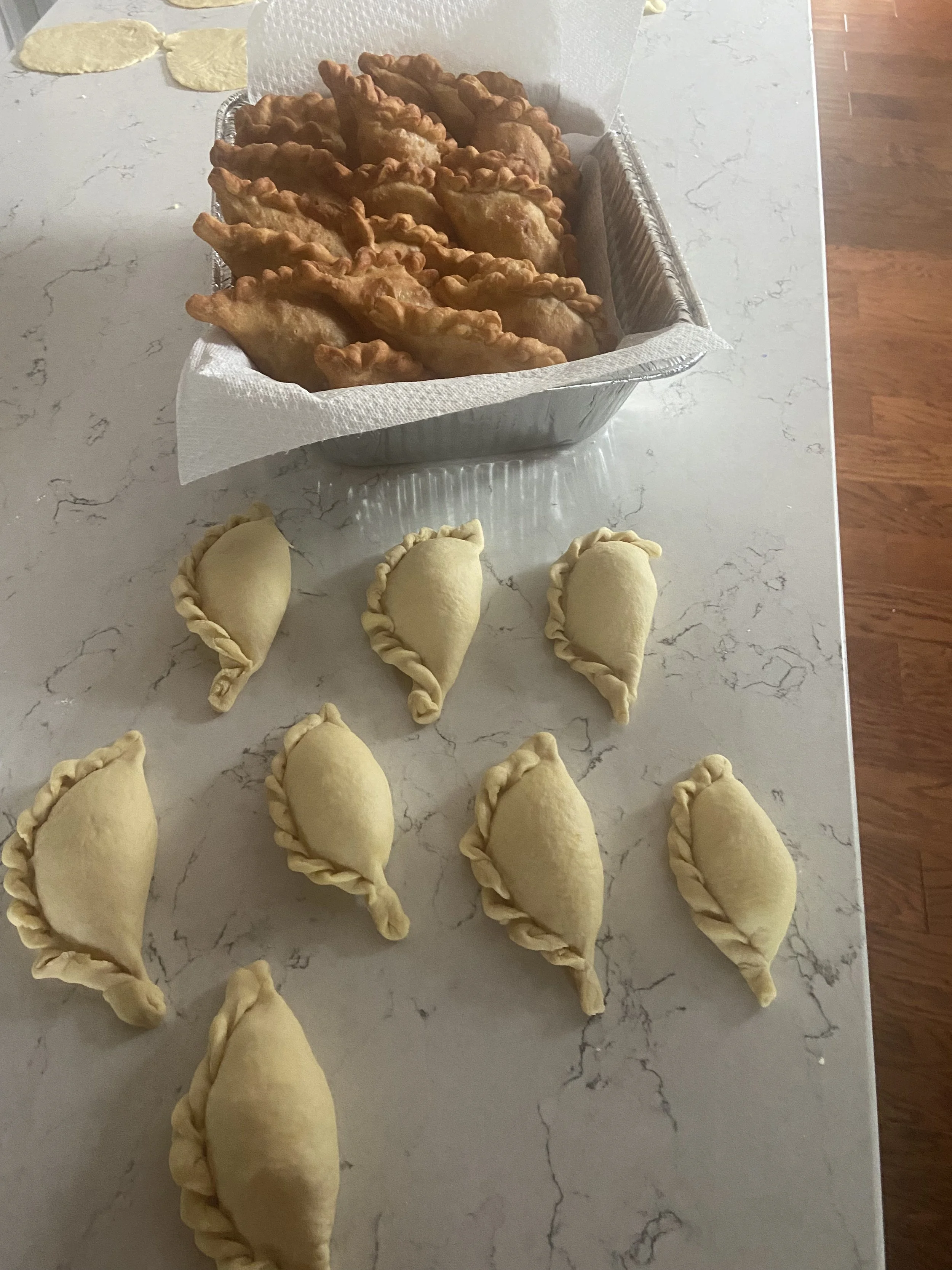 Unbaked empanadas lined up on a marble countertop with a basket of fried empanadas in the background.