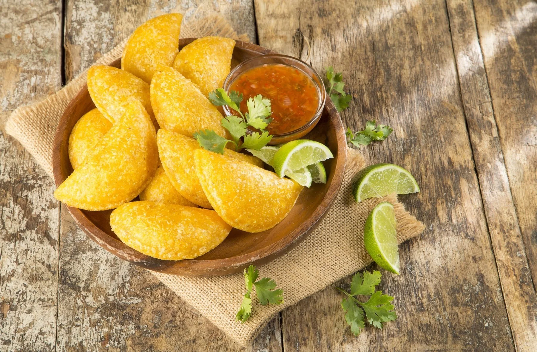 A wooden bowl filled with golden fried empanadas, garnished with lime wedges, cilantro, and served with side of salsa on a rustic wooden surface.