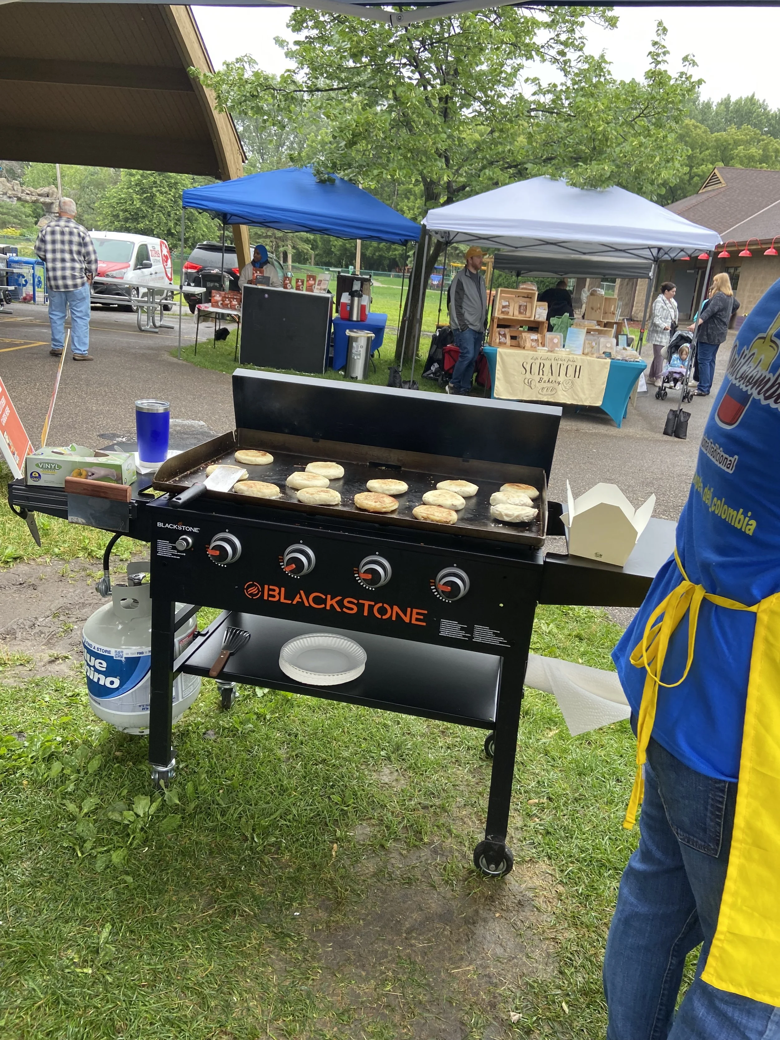 Grilled sausage patties cooking on a Blackstone grill at an outdoor market or fair with tents and people in the background.