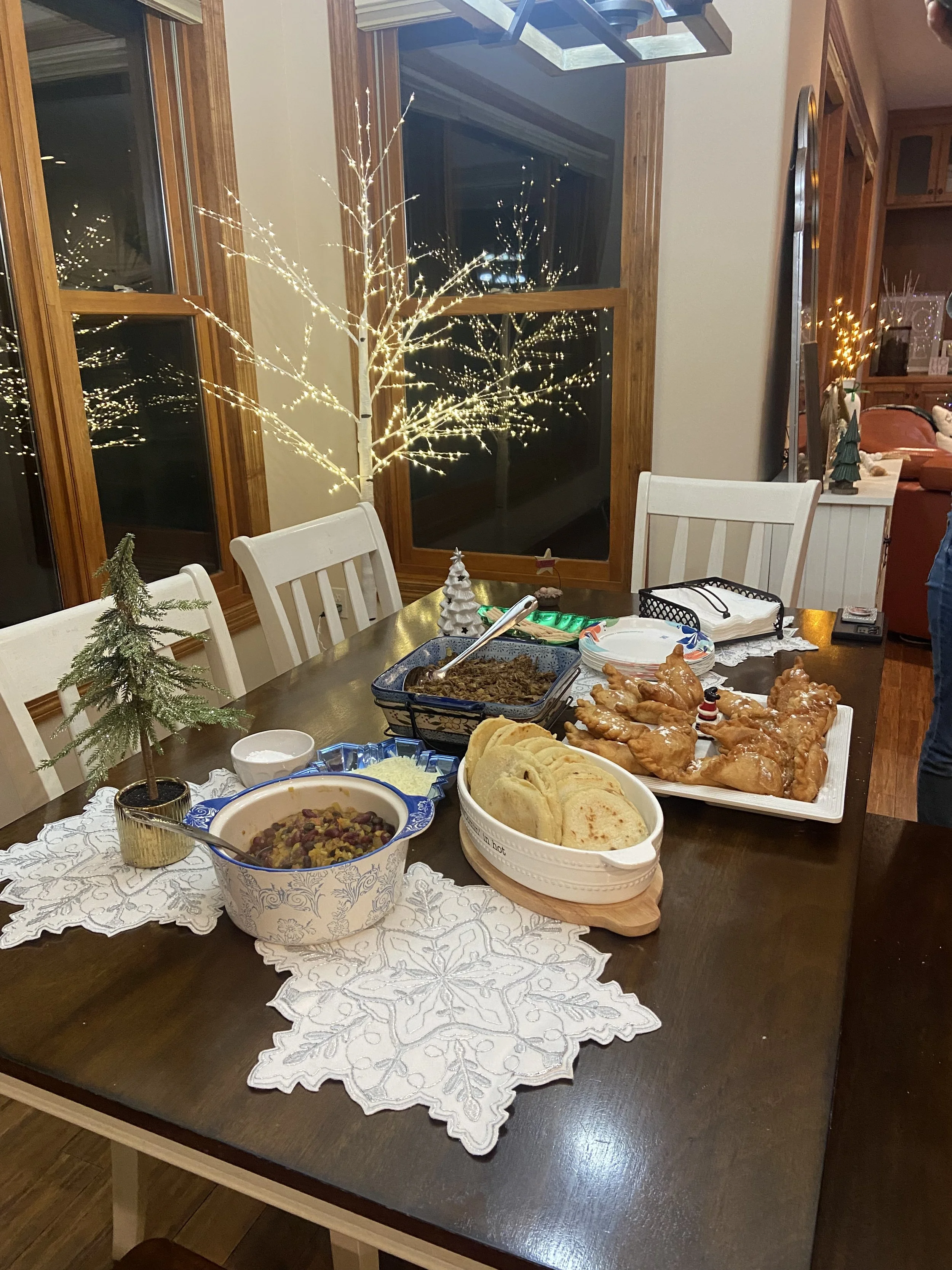 A holiday dinner table with various dishes including bread, a bowl of casserole, and chicken, decorated with Christmas trees and Christmas lights, with a snowy night scene outside the window.