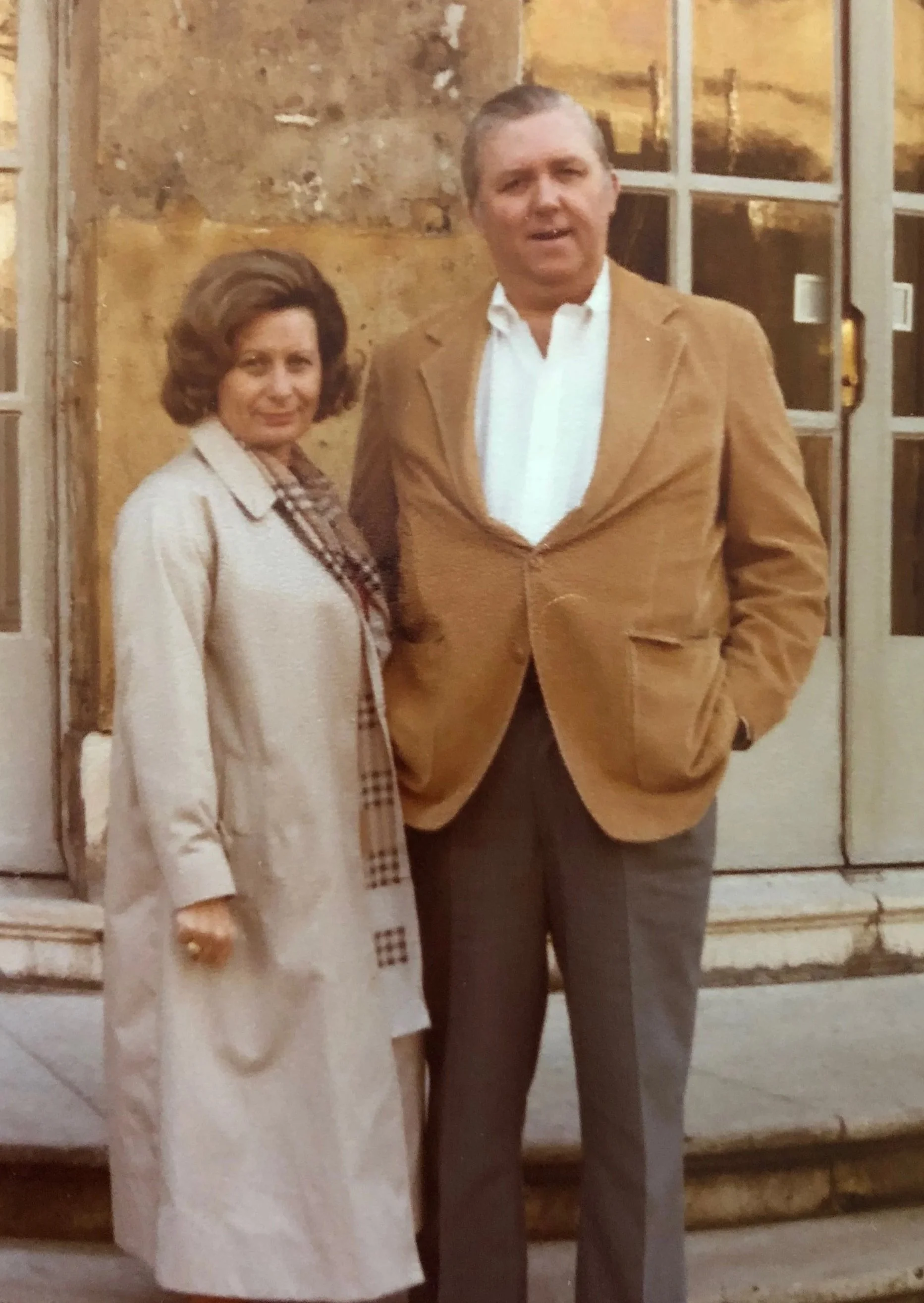 A man and a woman standing outside a building, dressed in vintage clothing from the mid-20th century.