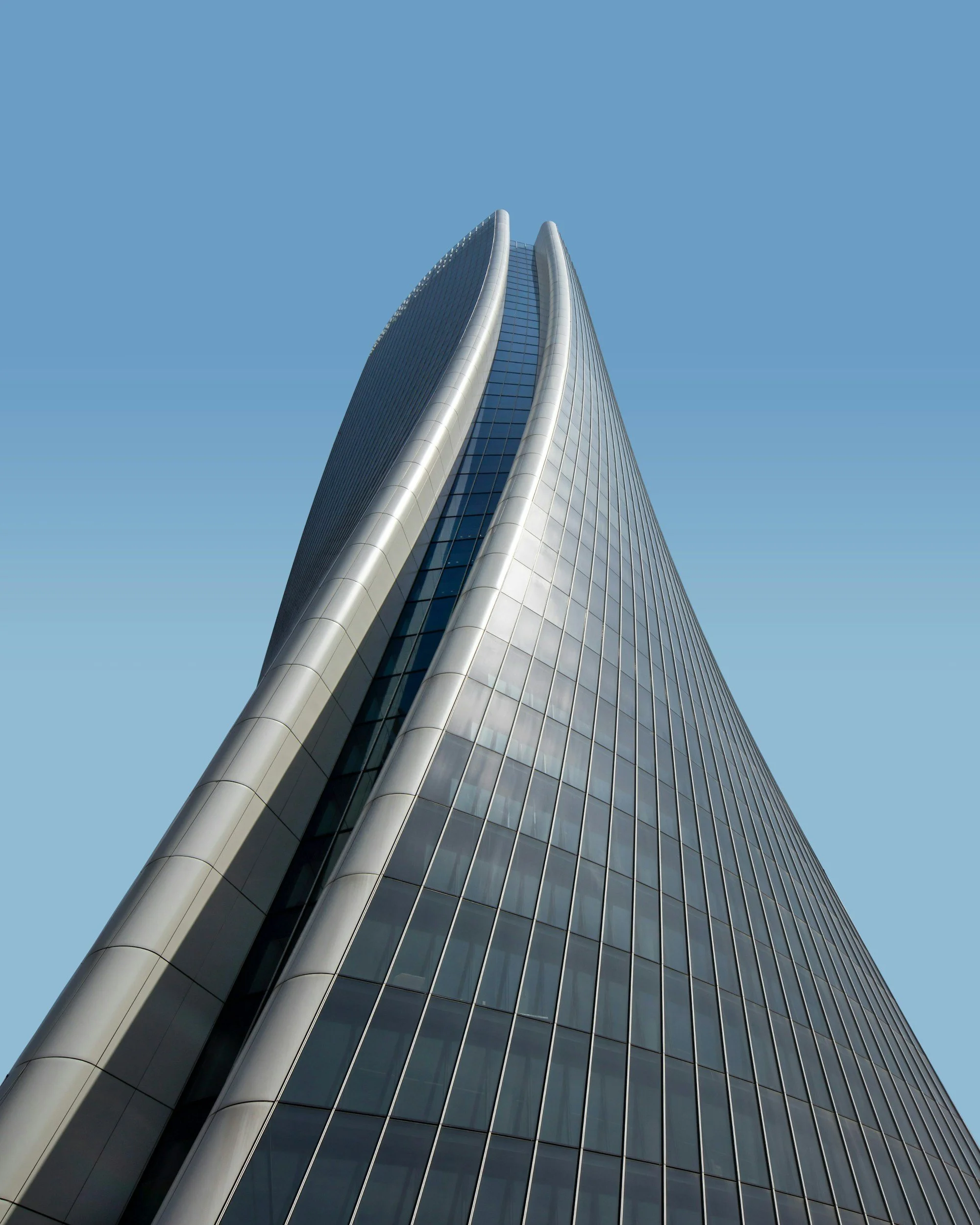 A tall modern skyscraper with reflective glass windows and rounded metal edges, seen from a low angle against a clear blue sky.