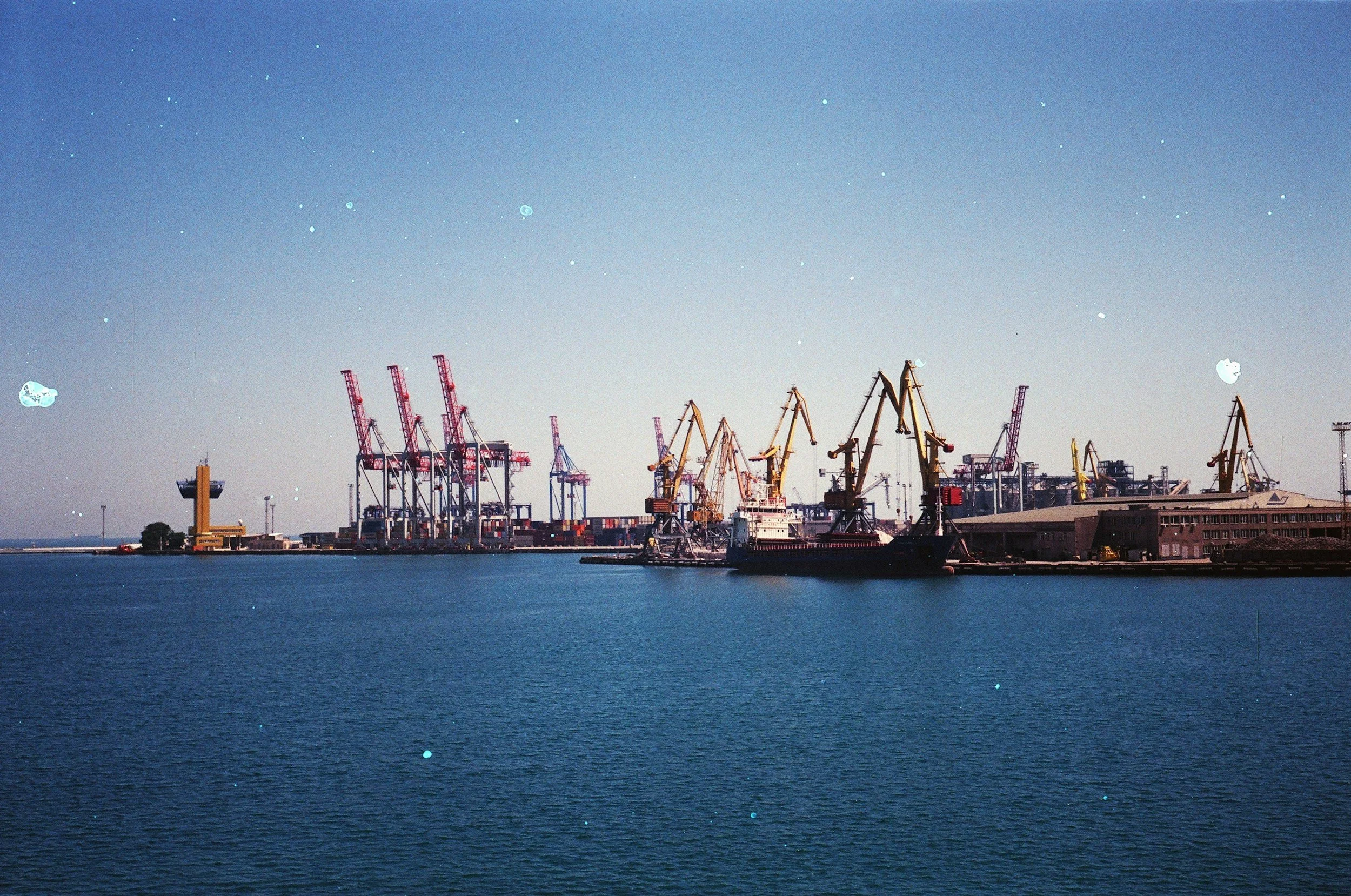 Port with multiple yellow and red cranes along the waterfront, with a body of water in the foreground and clear blue sky above.