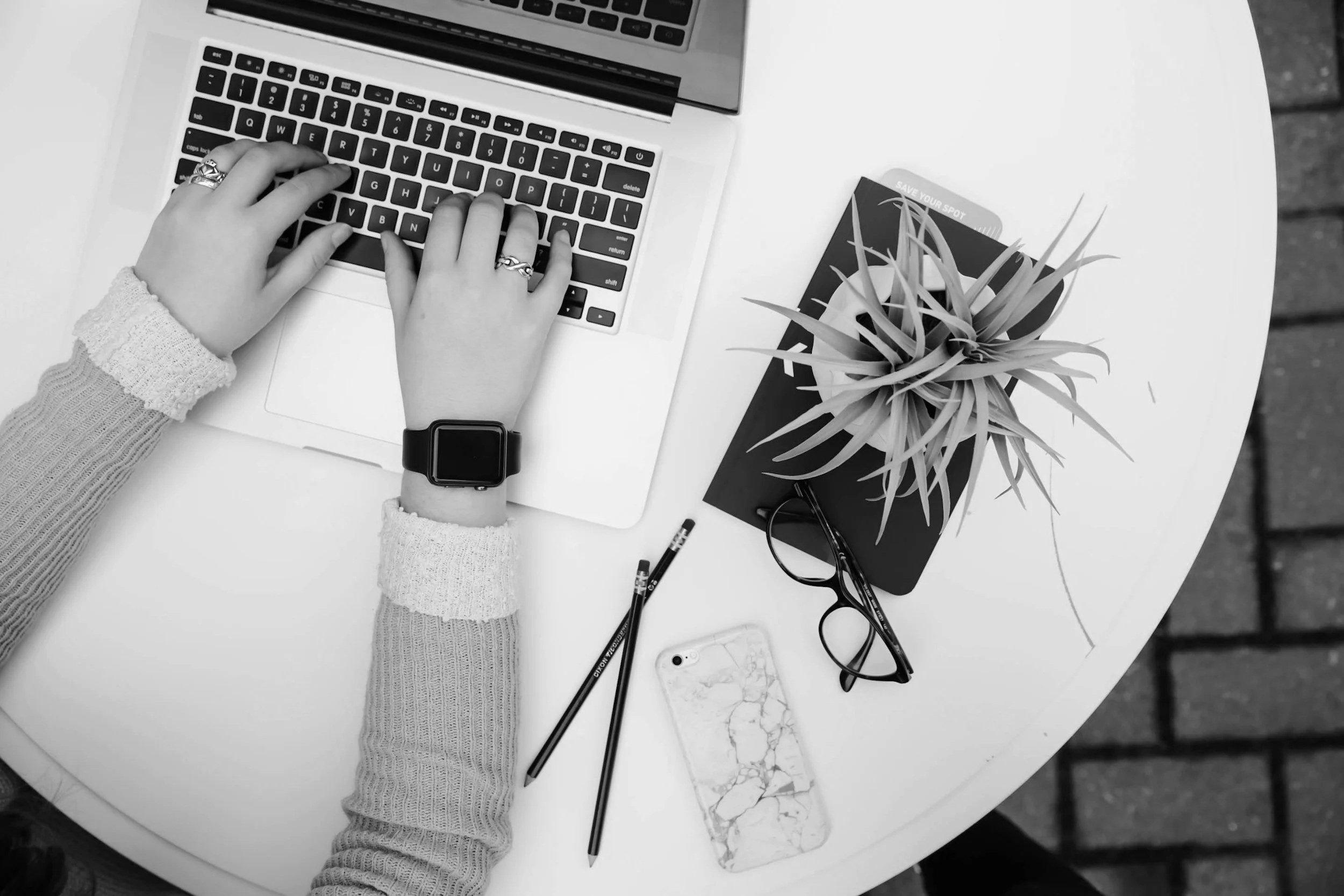 A top-down view of a workspace with a person typing on a laptop, wearing a sweater and a smartwatch, with a pair of glasses, a potted plant, a smartphone with a marble case, two pencils, and a notebook on a white table with a brick floor background.