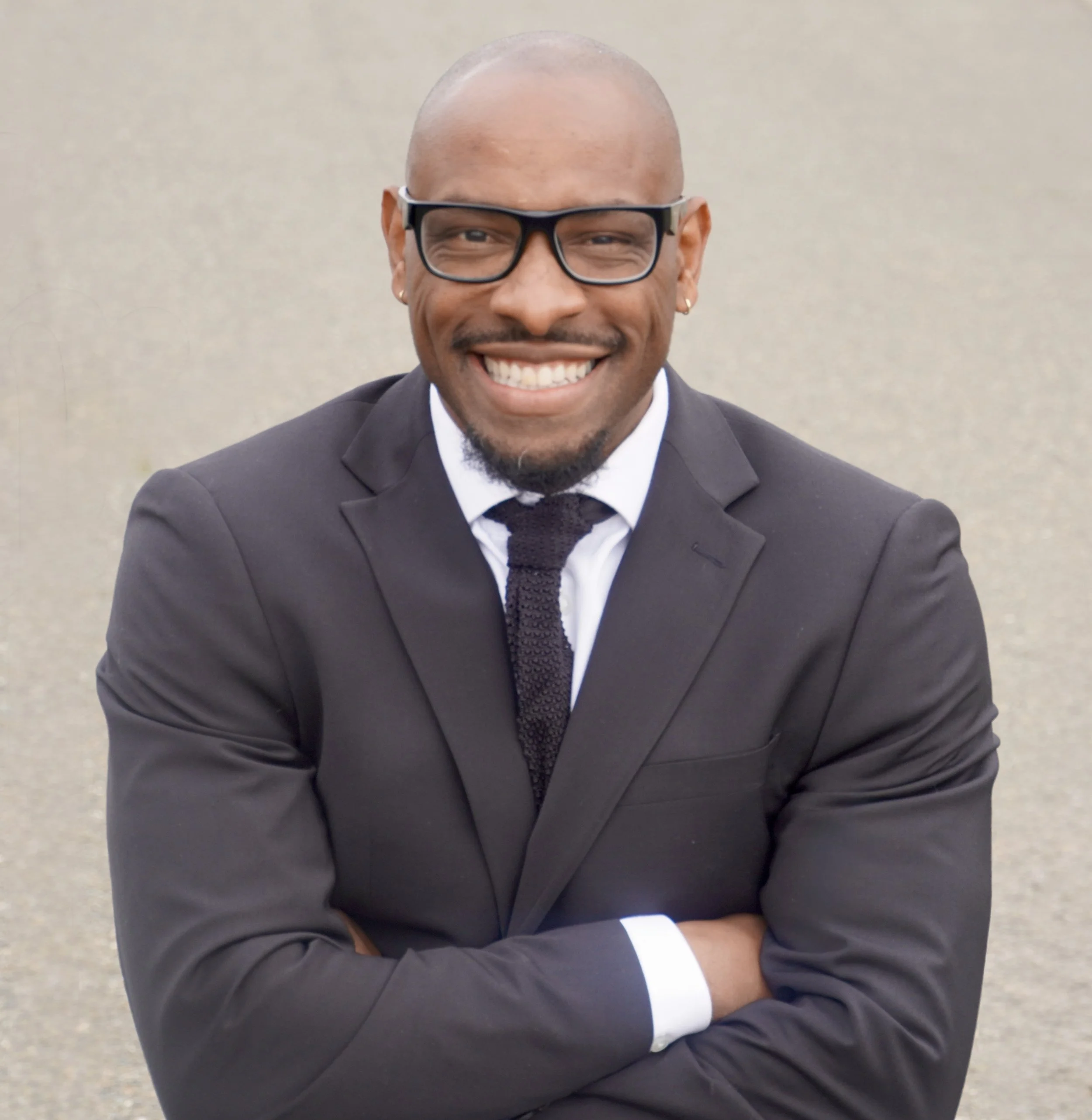 Smiling man in black suit, white shirt, black tie, glasses, standing outdoors with arms crossed.