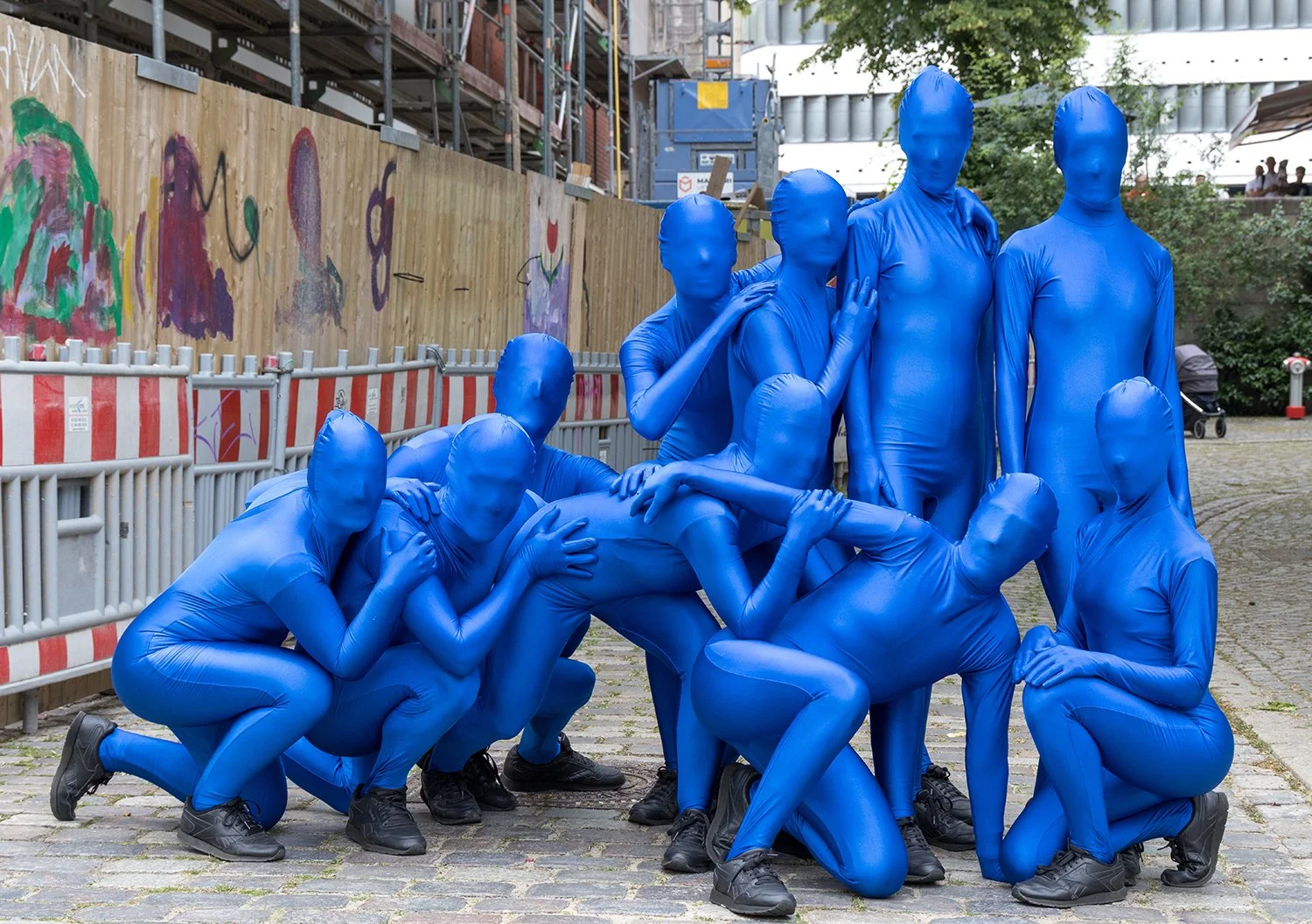 Mehrere Menschen in blauen, vollständigen Ganzkörperkostümen ohne Gesichtssicht posieren gemeinsam auf einer Straße neben einem Bauzaun und einem Gehweg.