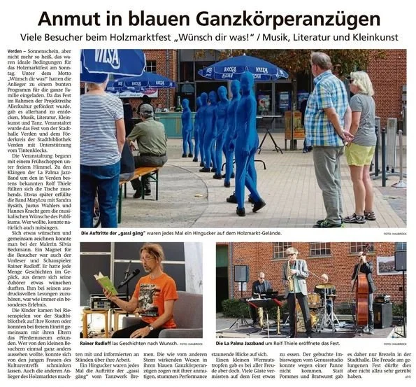 Bild zeigt einen öffentlichen Ort während eines Festes mit einer Aufführung. Im Vordergrund sind zwei Gruppen von Menschen: oben stehen Erwachsene, und unten sind Musiker mit Instrumenten. Im Hintergrund ist eine Reihe von blue ghost Kostümen, vermutlich als Teil der Aufführung.