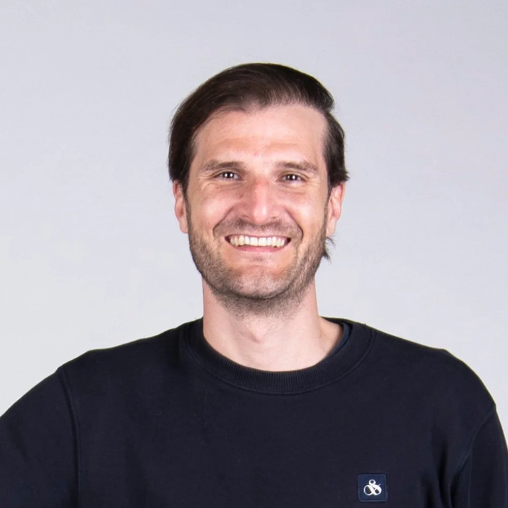 Lächelnder Mann mit dunklem Pullover vor neutralem Hintergrund.