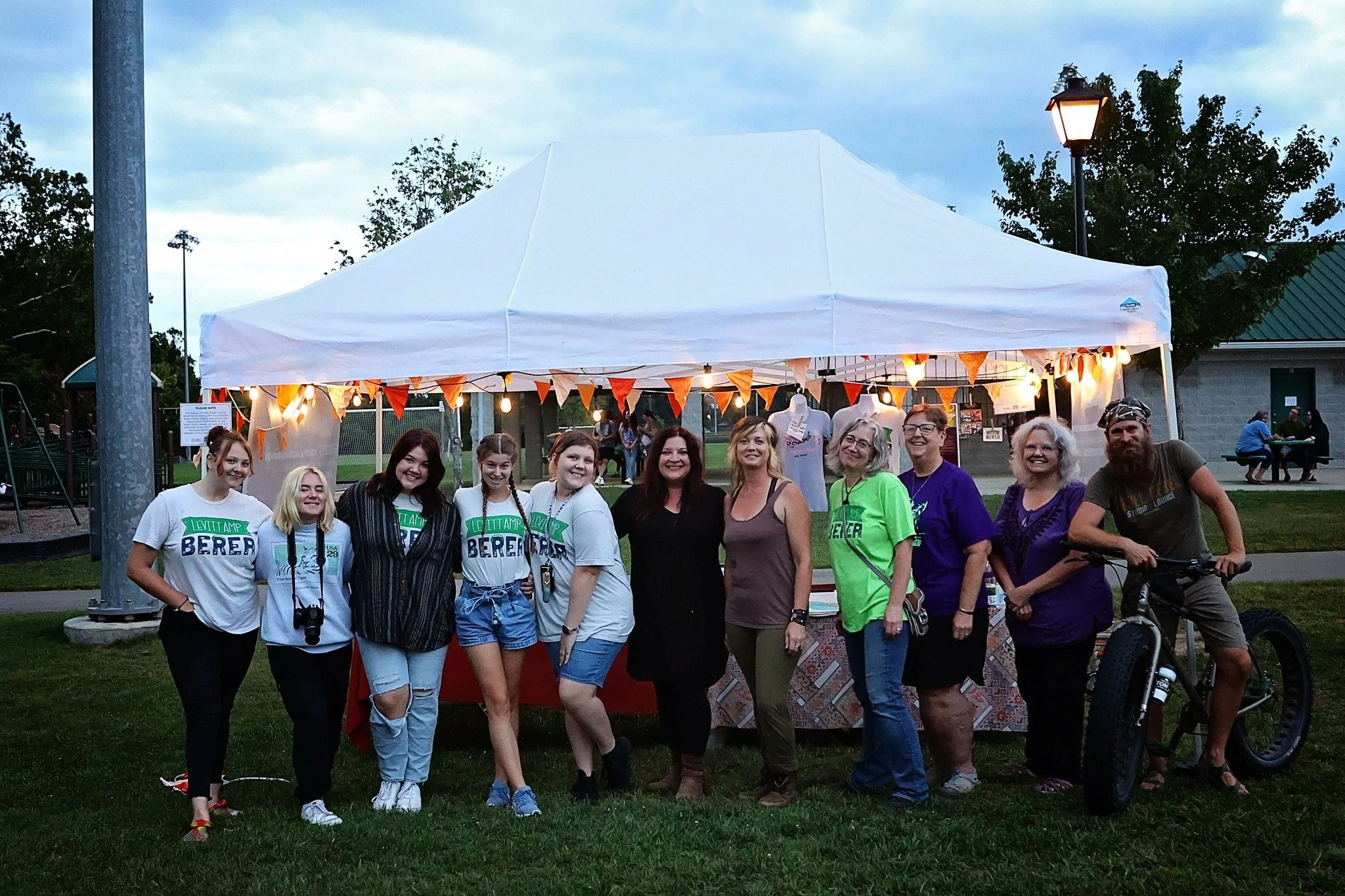 Group of people standing in front of a white canopy tent outdoors during evening, with some wearing event shirts, a bicycle, and a park with trees and benches in the background.