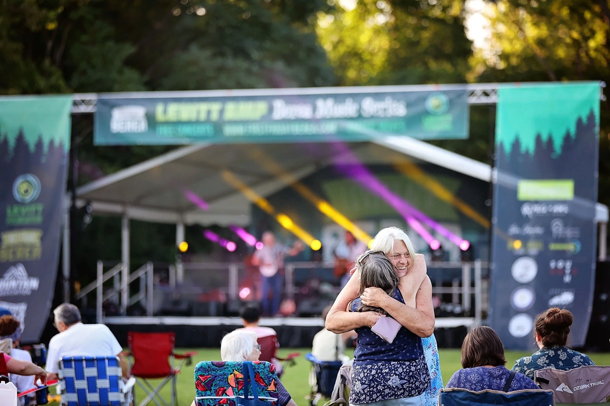 A group of people at an outdoor music concert, with a woman hugging another woman in the foreground, smiling. There is a band performing on a stage with colorful lighting in the background.