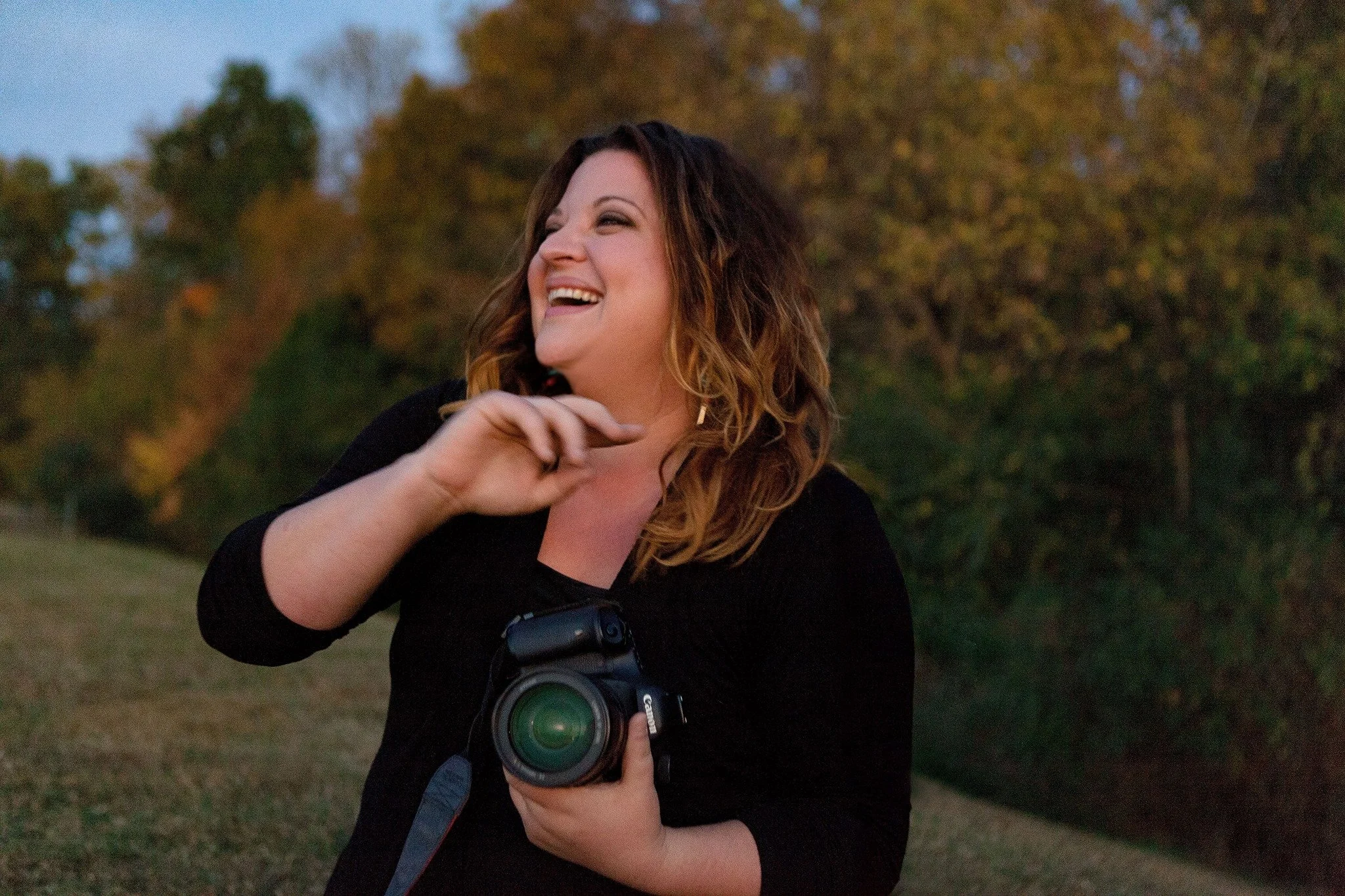 Erica Chambers, a woman with shoulder-length wavy hair smiling outdoors in a park during sunset, holding a Canon camera in her right hand, wearing a black long sleeve shirt.