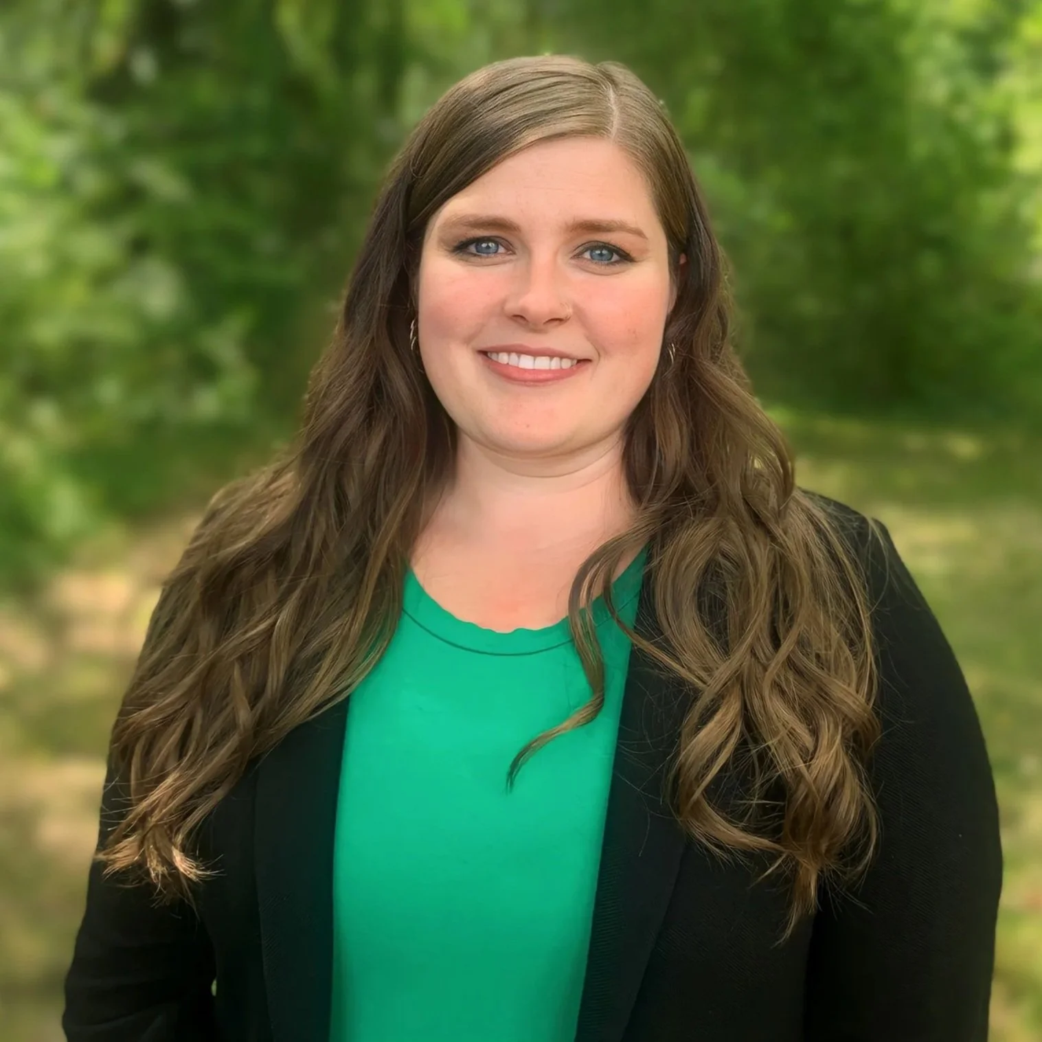 Samantha Dodson, a woman smiling outdoors with green trees in the background, wearing a black blazer and a green top.
