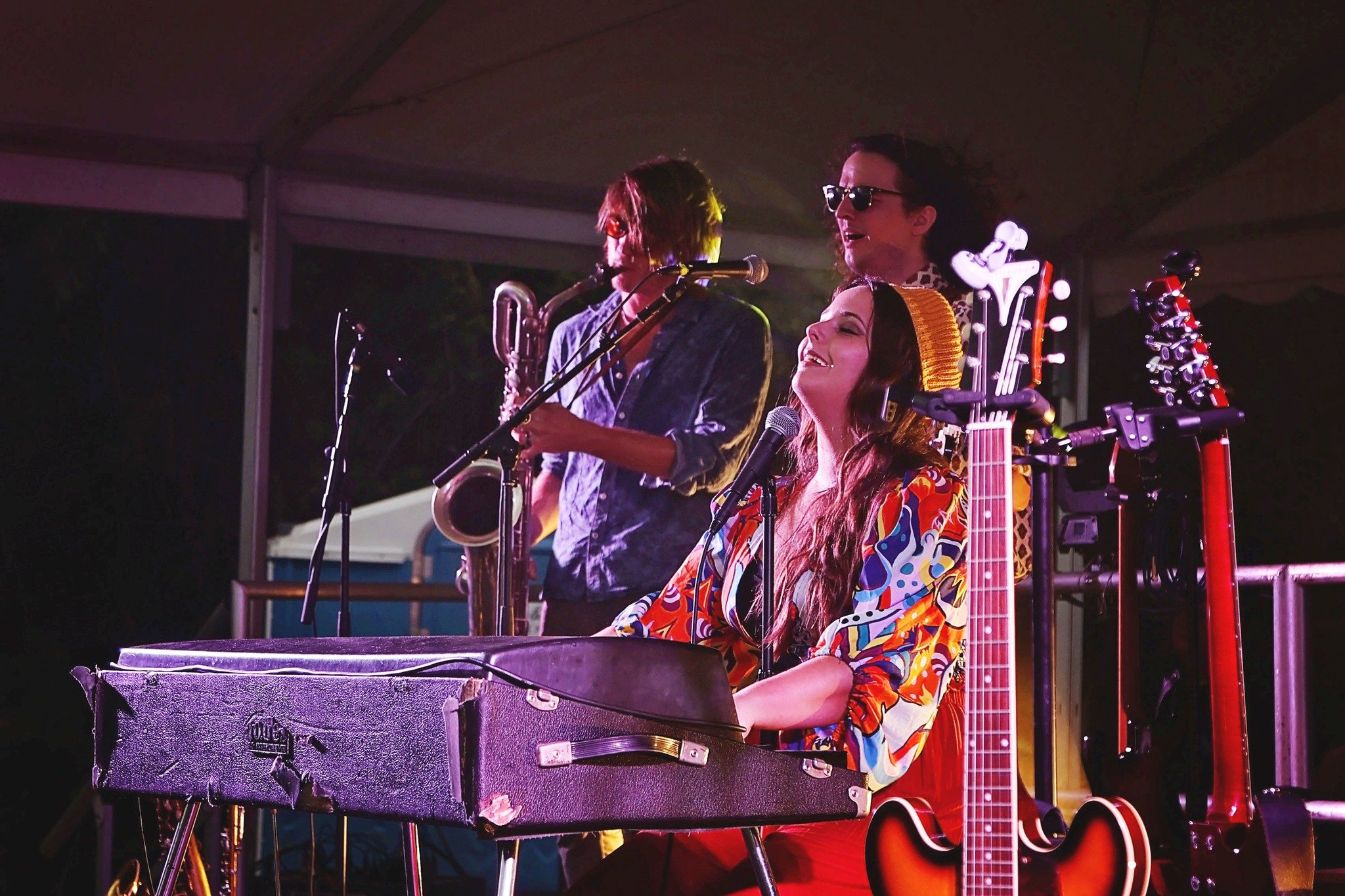 Musicians perform on stage, including Bee Taylor singing and playing keyboard, a saxophonist, and a person with an electric guitar.