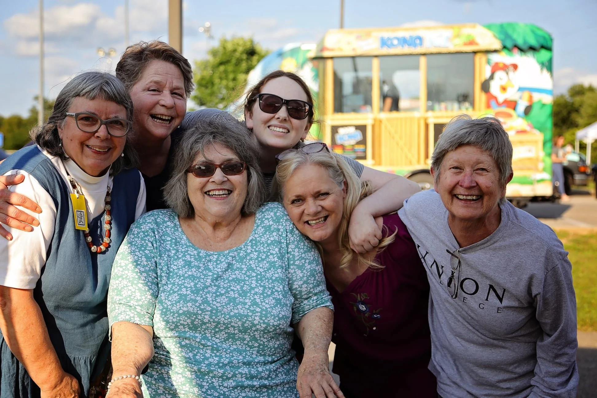 Seven women smiling and posing outdoors at a fair with a colorful food truck in the background.