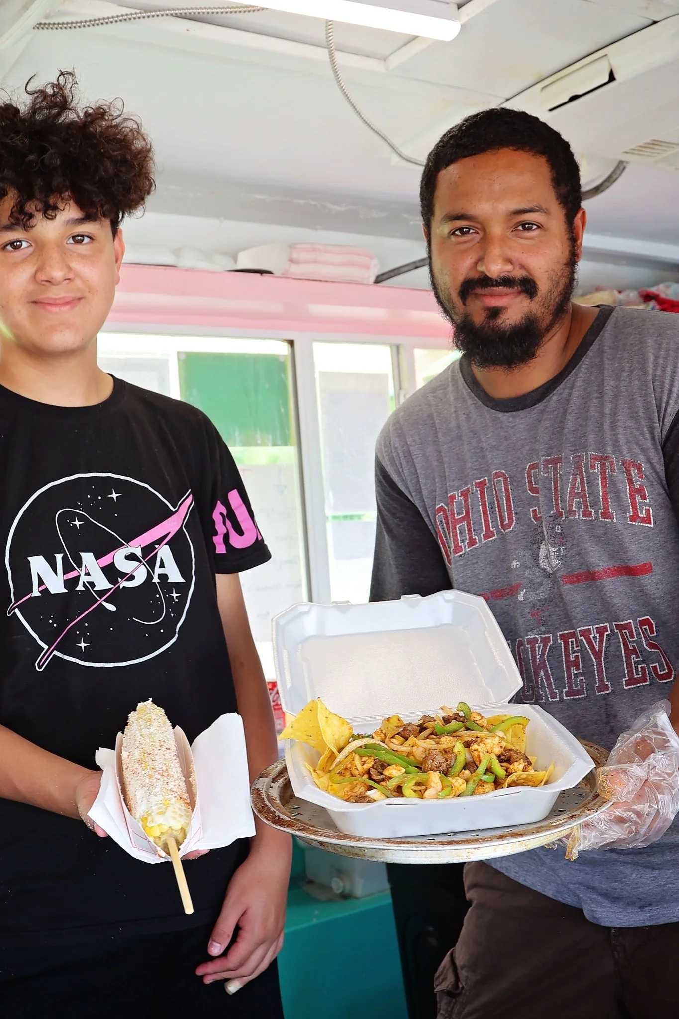 A young person holding a corn dog and an adult presenting a tray of loaded nachos at a food stand.