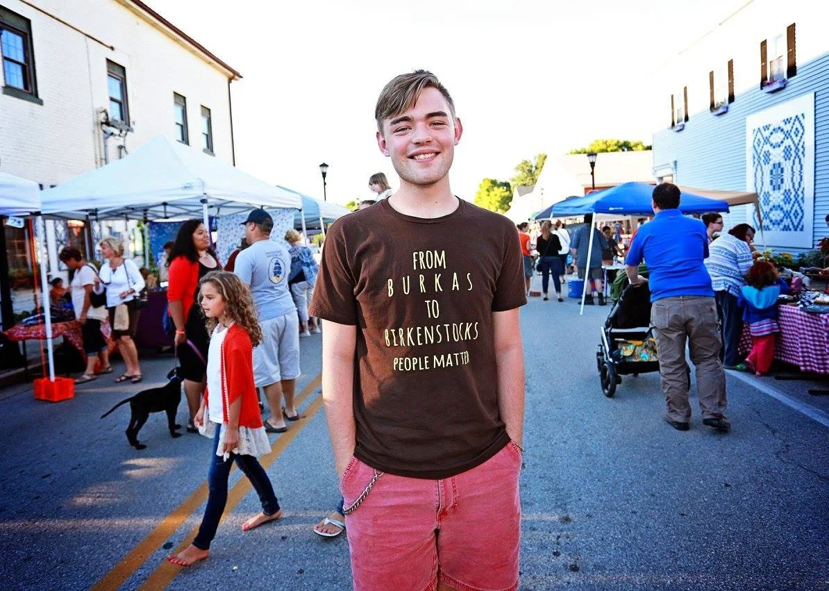 A young man wearing a brown T-shirt and pink shorts stands in front of a crowded street festival with people browsing tents and stalls, some with children and animals, against a backdrop of buildings and trees.