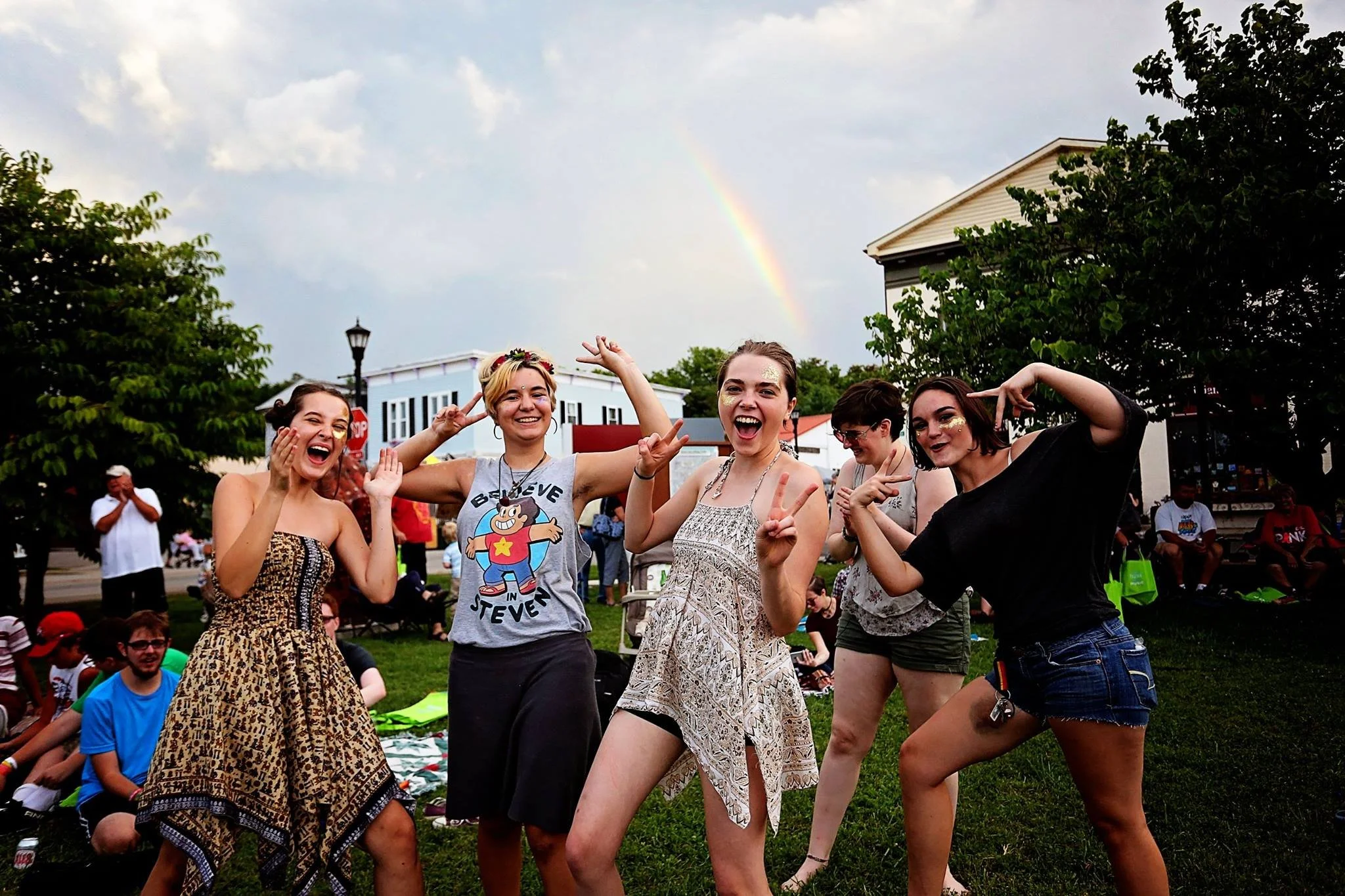 Group of five young women posing happily outdoors in front of a rainbow, with people sitting on the grass and trees in the background.