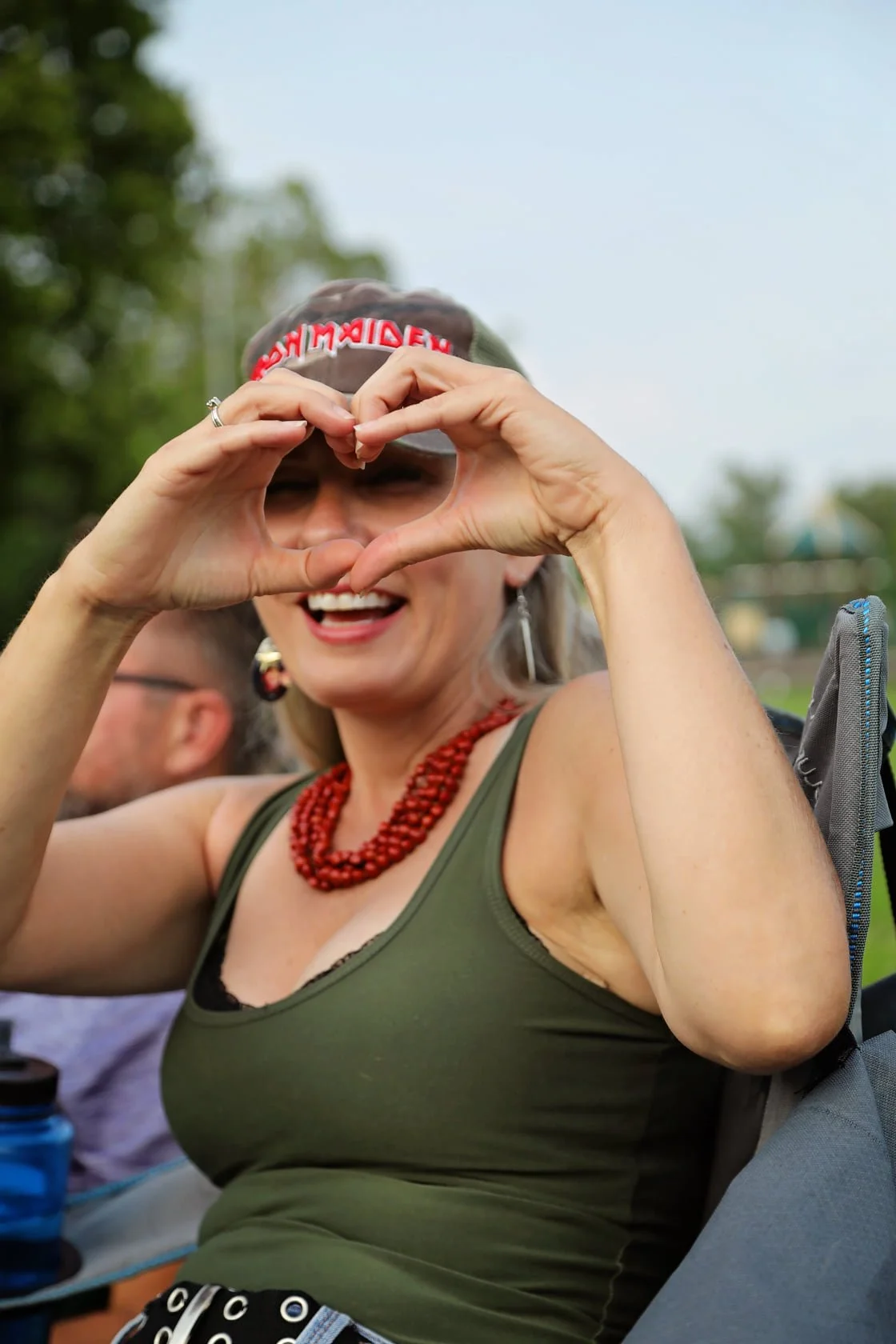 A smiling woman making a heart sign with her hands in front of her face, outdoors on a partly cloudy day, wearing a green top, red necklace, and a blackout Iron Maiden headband.