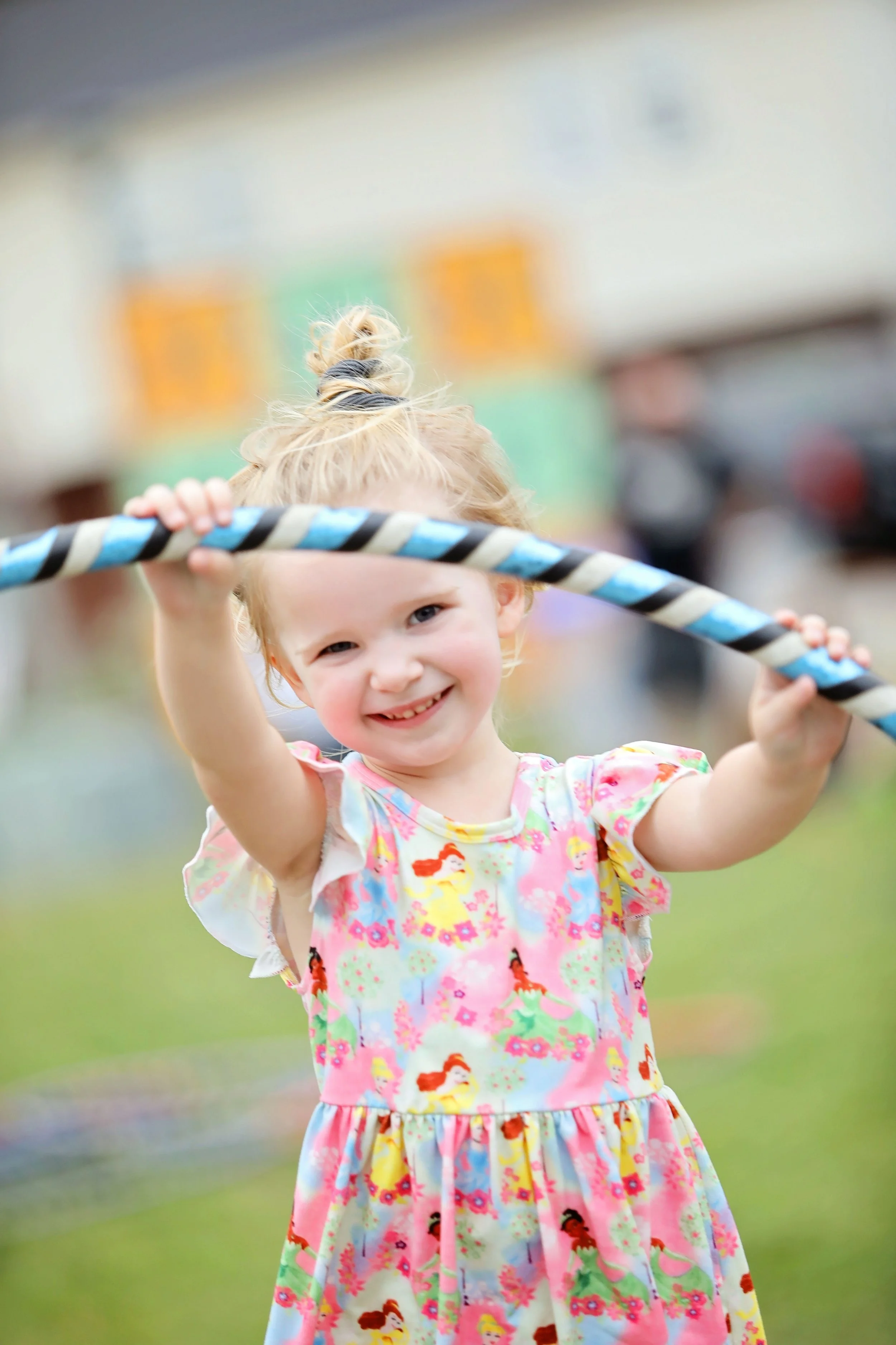 A young girl with blonde hair in a bun, smiling and playing with a hula hoop outdoors.