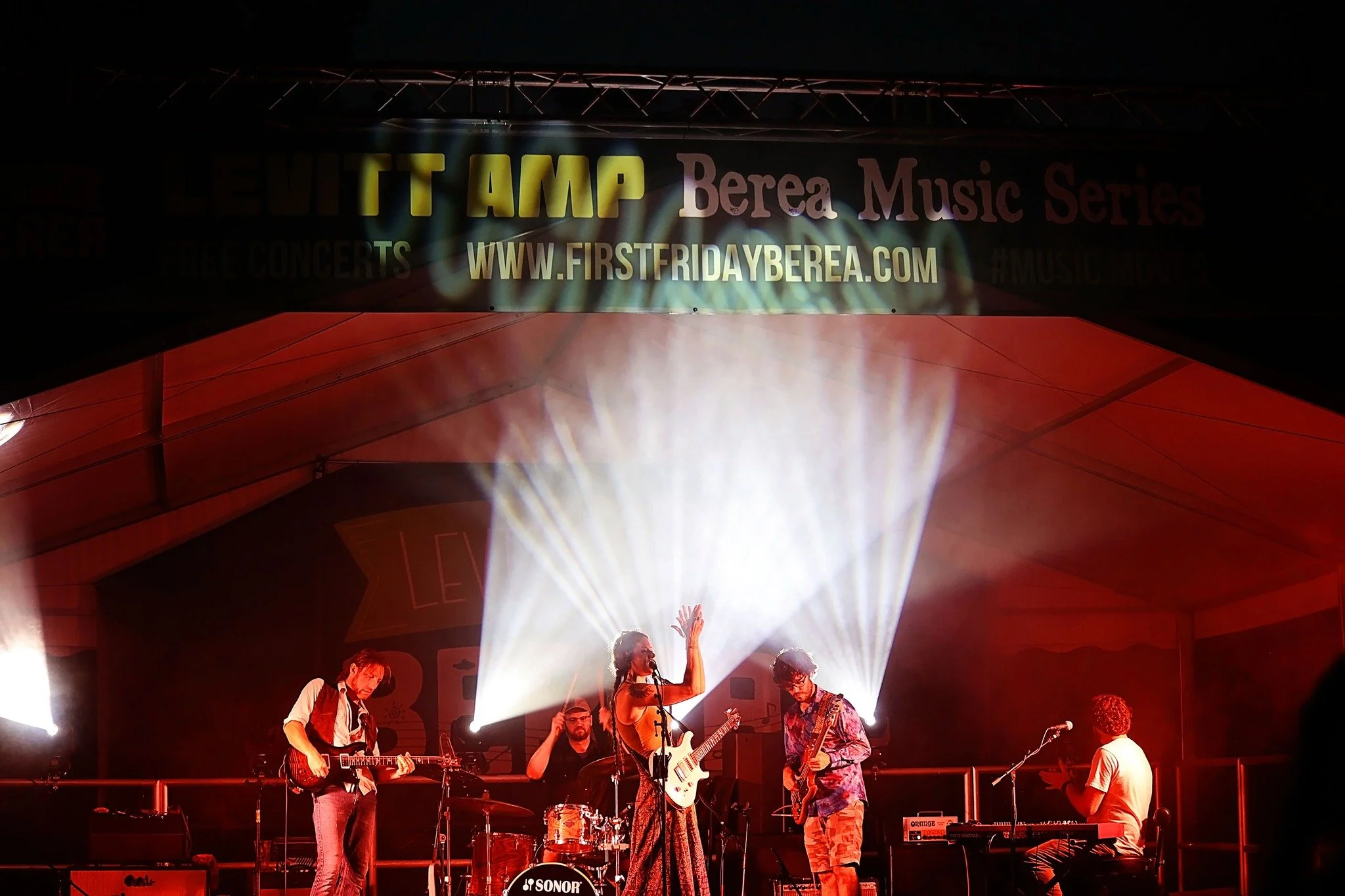 A band performing on stage at night with bright lights, including a guitarist, drummer, keyboardist, and a female singer, under a large sign advertising the Berea Music Series.