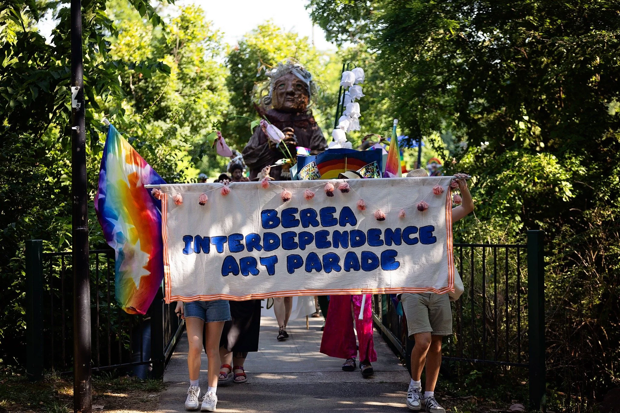 People participating in the BERSA Interdependence Art Parade, holding a large banner with rainbow flags, walking through a lush green park with trees.