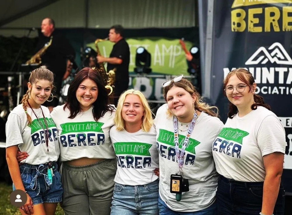 Five young women standing together at Leviitt Amp, wearing white T-shirts with a green banner and text, smiling at the camera, with a band playing in the background at an outdoor event.