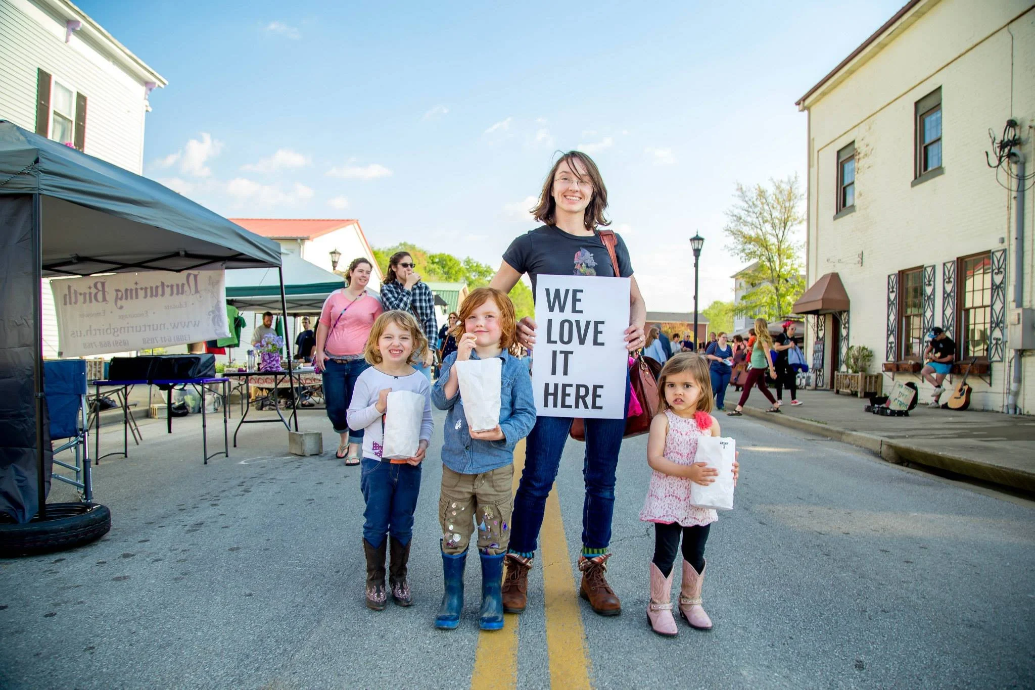 A woman and three young girls standing in the middle of a street during a community event. The woman holds a sign that says 'We Love It Here' and the girls hold paper bags. In the background, people are walking, and there are tents and buildings on either side of the street.