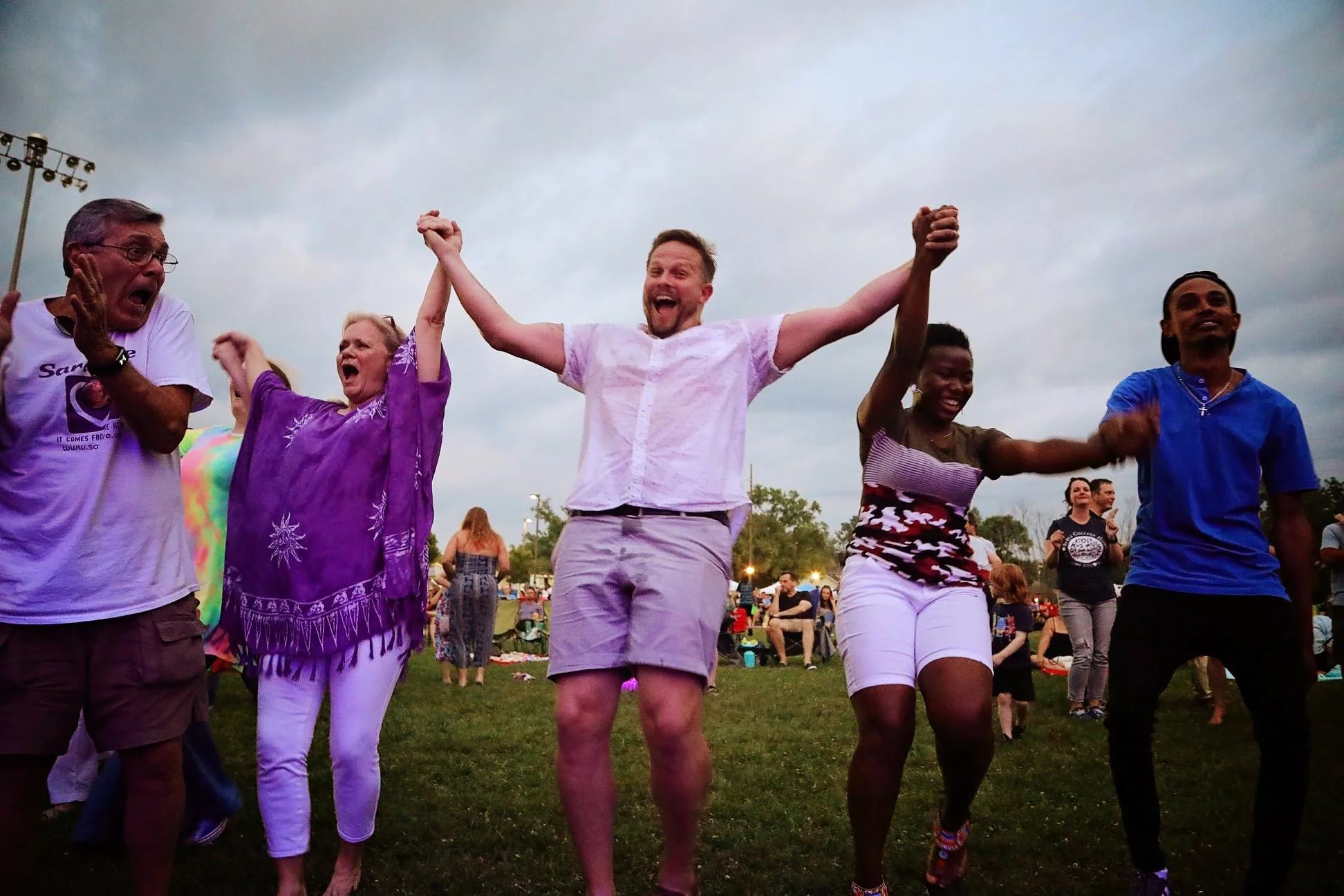 A diverse group of people holding hands and dancing outdoors at a festival or concert, with a cloudy sky overhead.