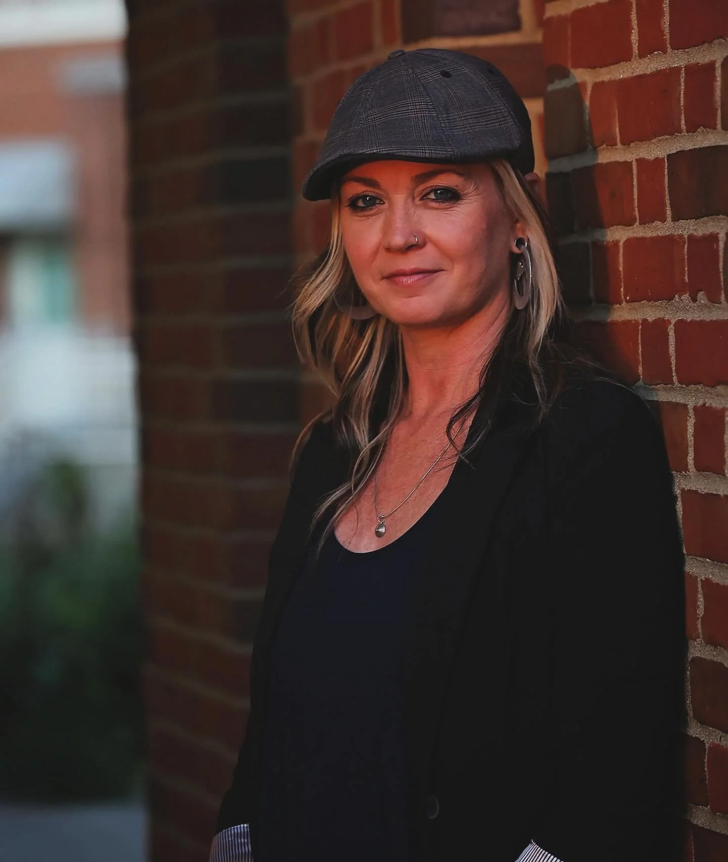 Ali Blair, a woman with blonde hair, wearing a gray flat cap, black top, and black blazer, standing against a brick wall outdoors.
