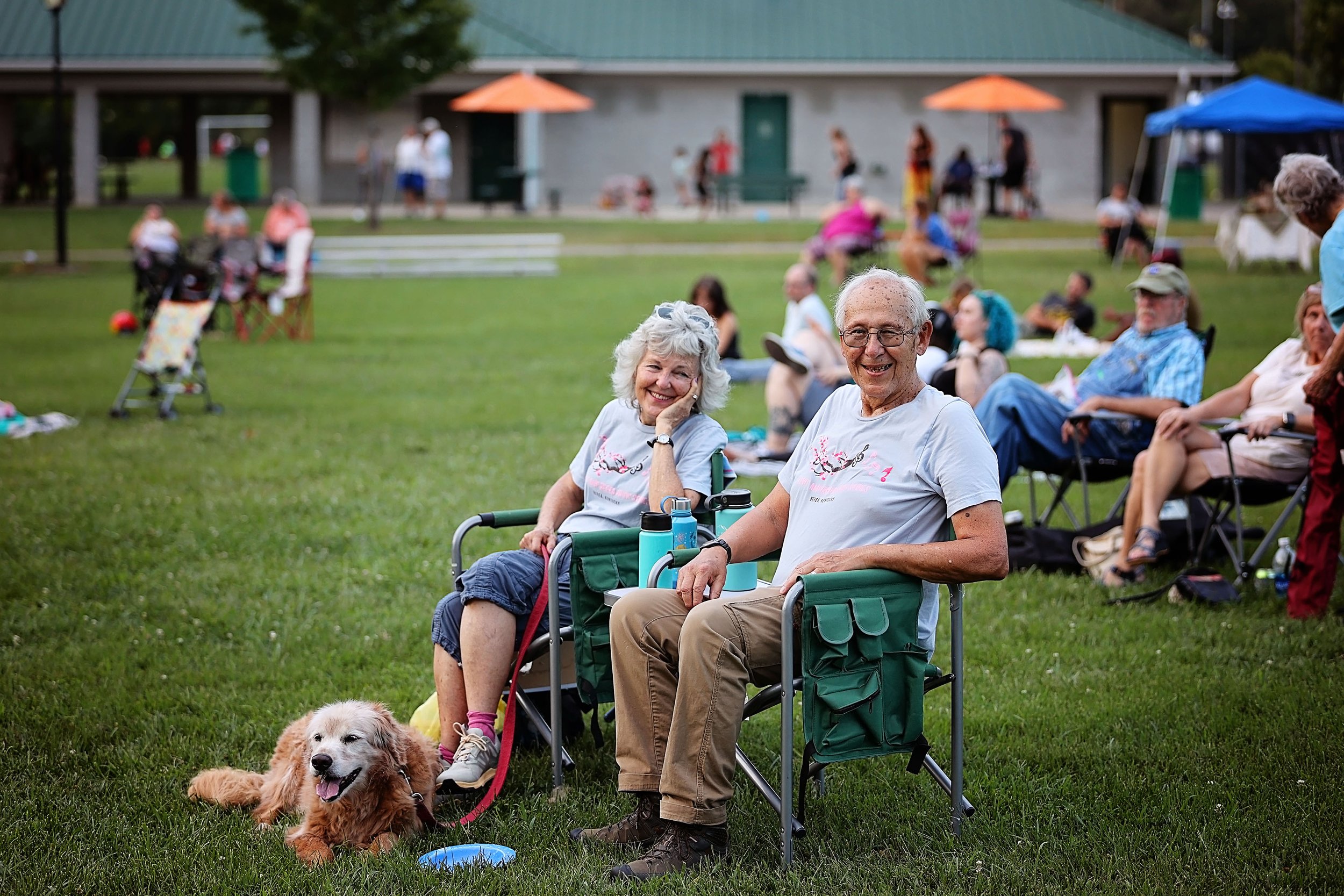 Smiling elderly couple sitting in camping chairs on a grassy field with a golden retriever dog, surrounded by people at an outdoor gathering with tents and umbrellas in the background.