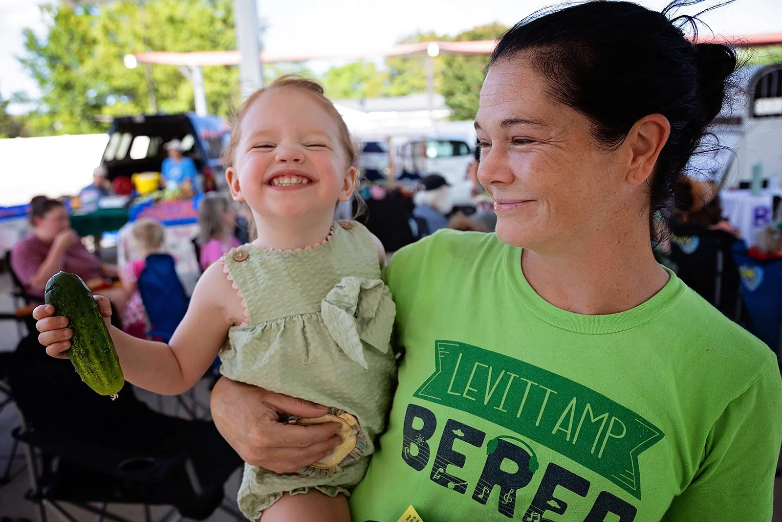 A woman holding a smiling young girl at an outdoor event, with the girl holding a cucumber. The woman wears a bright green T-shirt with 'Levitttamp Beers' written on it, and there are other people and food stands in the background.