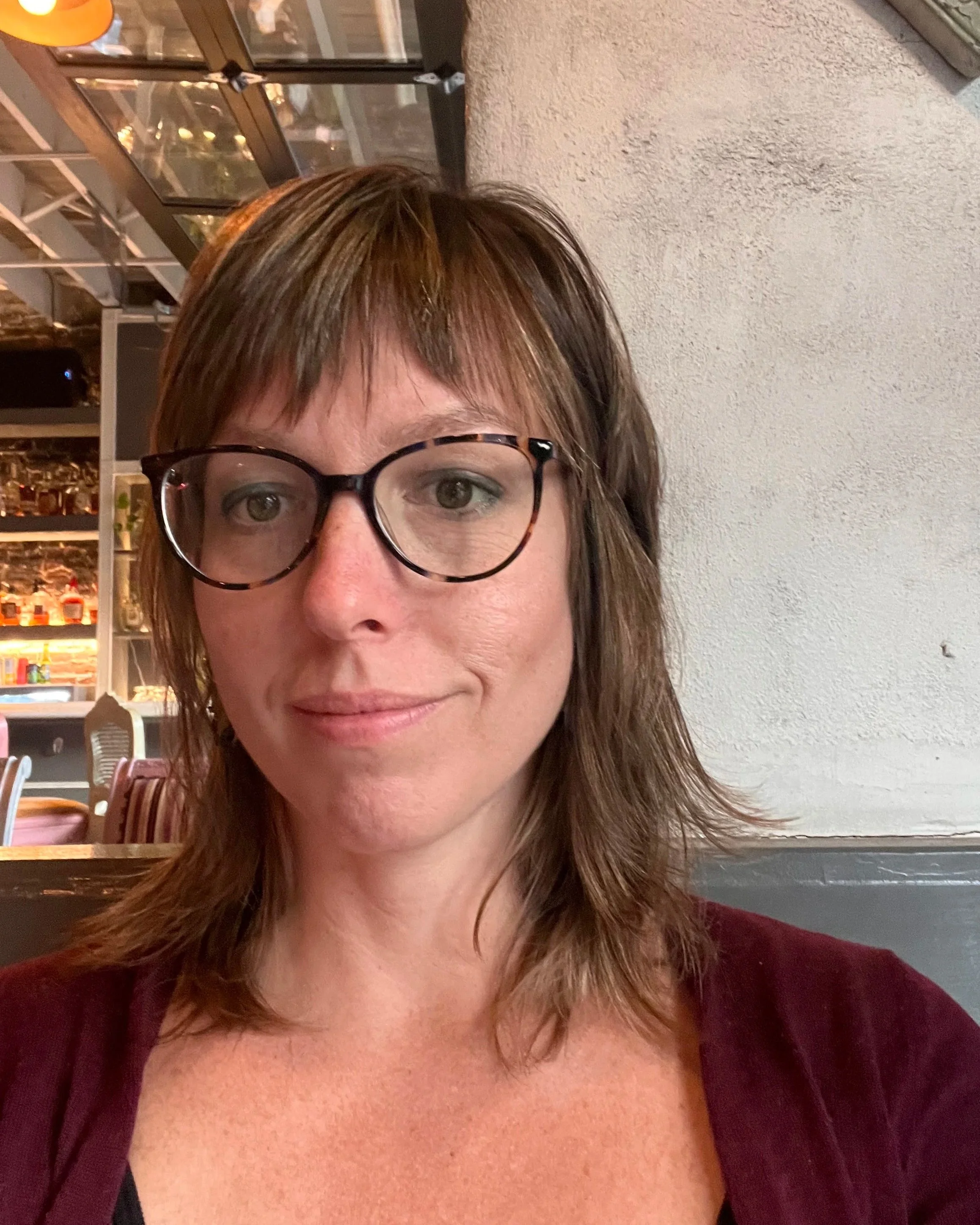 A woman with brown hair, glasses, and light makeup smiling in a restaurant or cafe setting with shelves of bottles and glasses in the background.