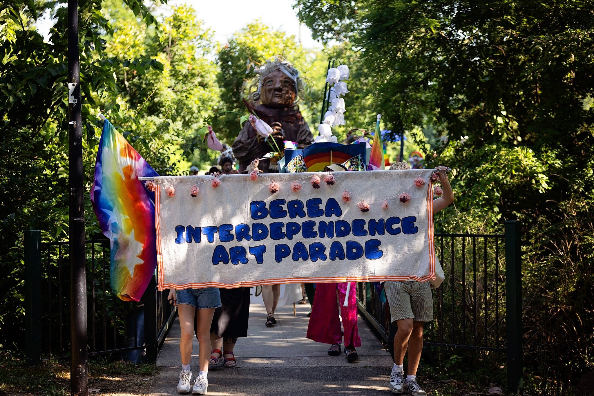 People holding a banner that reads 'BERSA INDEPENDENCE ART PARADE' during a parade through a park with trees.