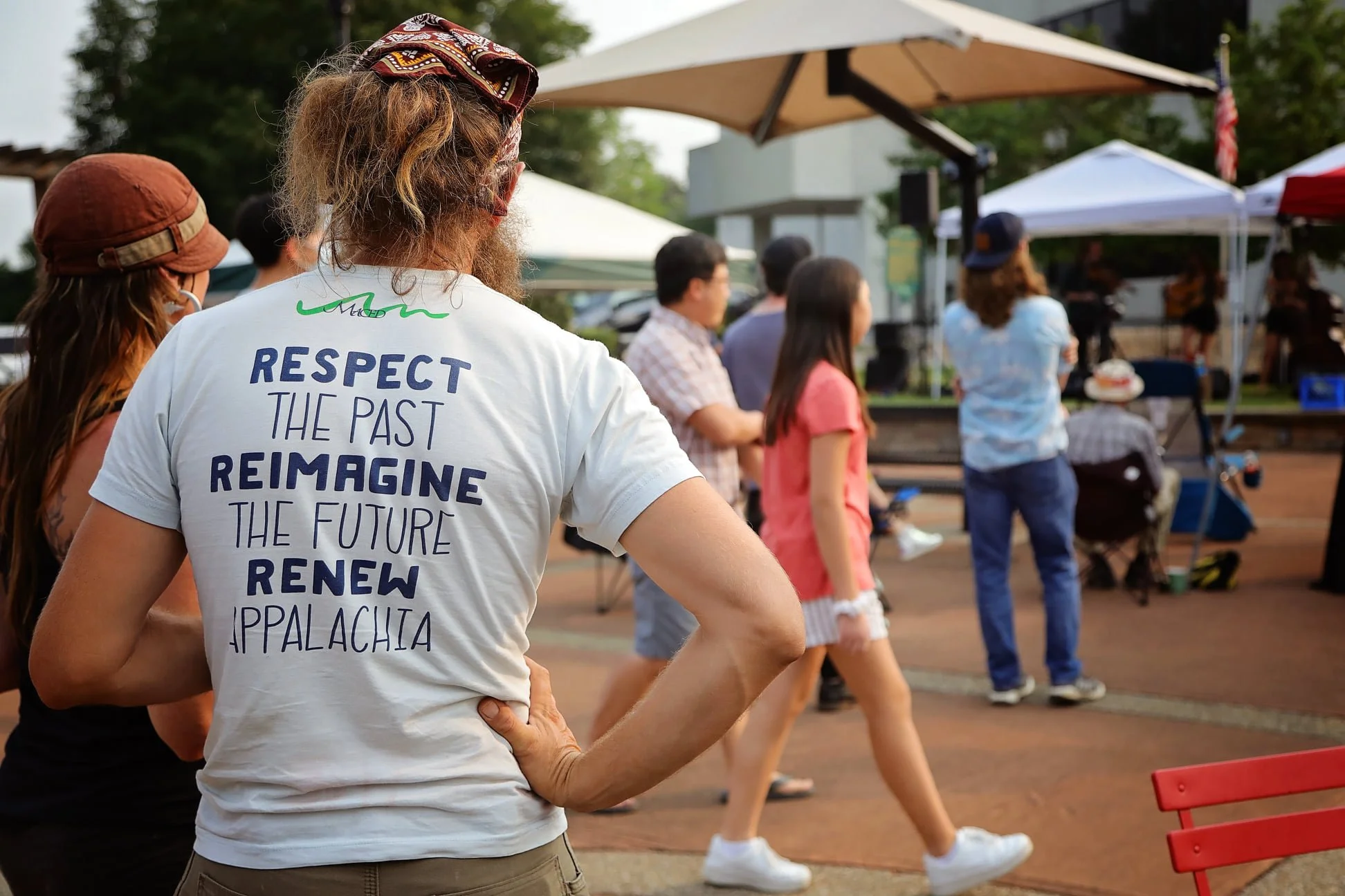 People attending an outdoor community event with tents, a stage, and a speaker, with a man in a white t-shirt that reads, 'Respect the past, reimagine the future, renew Appalachia.'